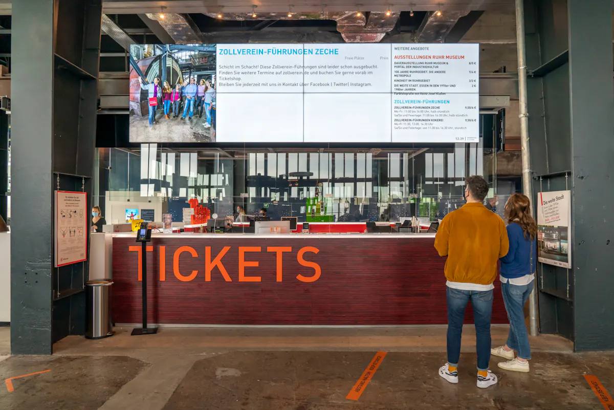 Zwei Personen stehen vor einem modernen Ticketschalter mit der Aufschrift TICKETS in großen orangefarbenen Buchstaben. Über dem Schalter hängt ein großer Bildschirm, der Informationen zu Führungen und Ausstellungen auf dem Gelände der Zeche Zollverein anzeigt. Auf dem Bildschirm sind unter anderem Zollverein-Führungen, Preise und Zeiten zu sehen sowie ein Foto einer Besuchergruppe im Bergwerk. Hinter dem Schalter befinden sich Mitarbeiterinnen und Mitarbeiter in einem hellen, gläsernen Empfangsbereich. Die Umgebung wirkt industriell und modern gestaltet, mit Sichtbeton und offenen Stahlträgern. Links und rechts hängen zusätzliche Infotafeln an den Pfeilern.