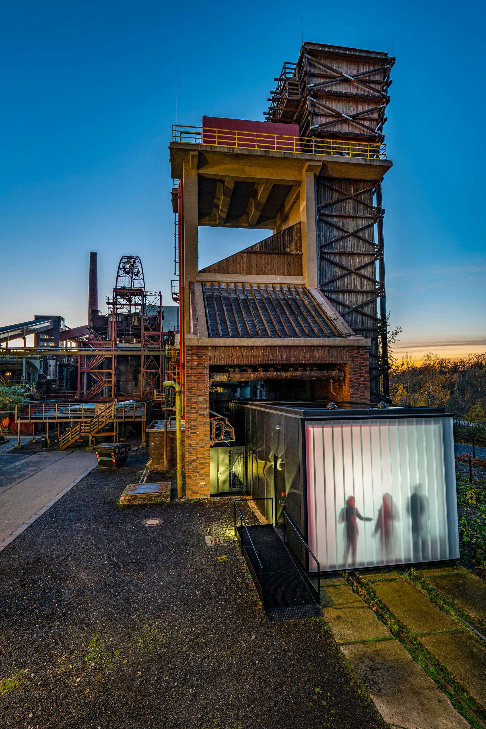Außenaufnahme eines ehemaligen Industriekomplexes bei Sonnenuntergang mit einer imposanten Struktur aus Beton, Stahl und Backstein. Im Vordergrund rechts befindet sich ein moderner, quaderförmiger Glasbau mit transluzenter Fassade, hinter der die Silhouetten von drei Personen im Inneren zu erkennen sind. Die Szene kombiniert historische Industriearchitektur mit zeitgenössischem Ausstellungs- oder Eingangsdesign. Die Lichtstimmung ist warm und kontrastiert mit dem kühlen Blau des Abendhimmels. Links im Hintergrund sind weitere Industrieruinen, eine Schornsteinanlage und begehbare Stege sichtbar.