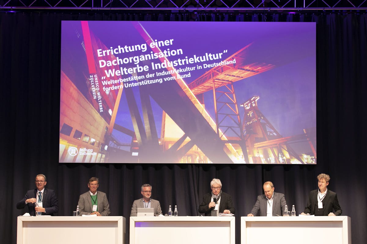 Sechs Männer sitzen und stehen hinter weißen Pulten auf einer Bühne vor einem großen, beleuchteten Hintergrundbild, das ein Industriebauwerk in Abendstimmung zeigt. Auf dem Hintergrund steht in weißer Schrift: Errichtung einer Dachorganisation Welterbe Industriekultur – Welterbestätten der Industriekultur in Deutschland fordern Unterstützung vom Bund. Links unten auf dem Bild ist das Logo der Stiftung Zollverein zu sehen. Die Männer tragen Anzüge oder Jacketts und Namensschilder. Die Atmosphäre ist formell, wie bei einer Konferenz oder Podiumsdiskussion. Die Bühne ist von schwarzen Vorhängen umgeben, der Raum ist technisch ausgeleuchtet.