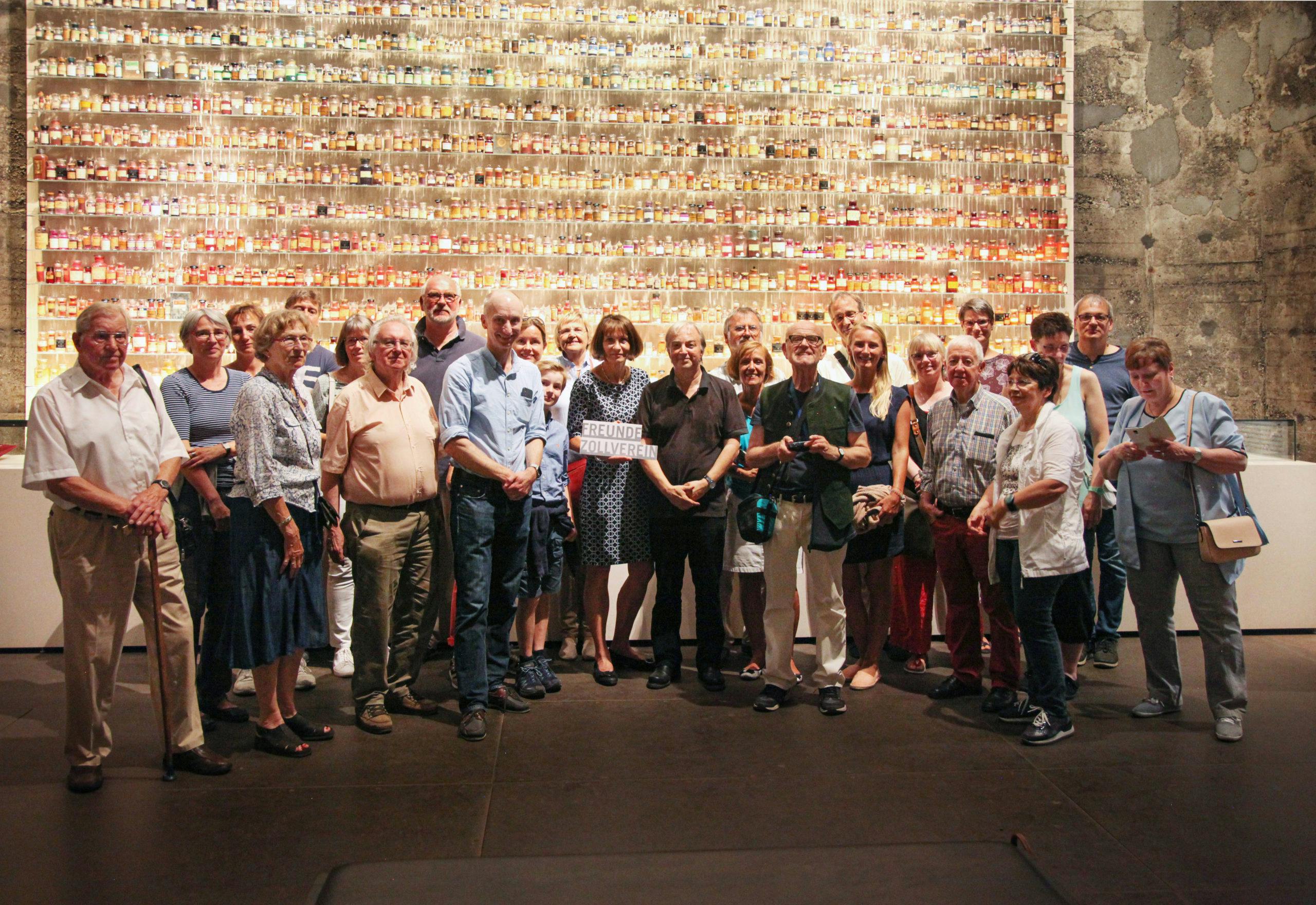 Gruppenfoto von etwa 25 Personen unterschiedlichen Alters in einem Ausstellungsraum mit industriellem Ambiente. Die Gruppe steht vor einer großformatigen Wandinstallation aus unzähligen kleinen bunten Glasfläschchen, die in Reihen angeordnet und beleuchtet sind. In der Mitte des Bildes hält eine Frau ein Schild mit der Aufschrift Freunde Zollverein. Die Menschen blicken lächelnd in die Kamera, einige stehen eng beieinander, andere blättern in Papieren oder unterhalten sich. Die Szene vermittelt Gemeinschaft, Interesse an Kunst und Kultur und Zugehörigkeit zur Institution.