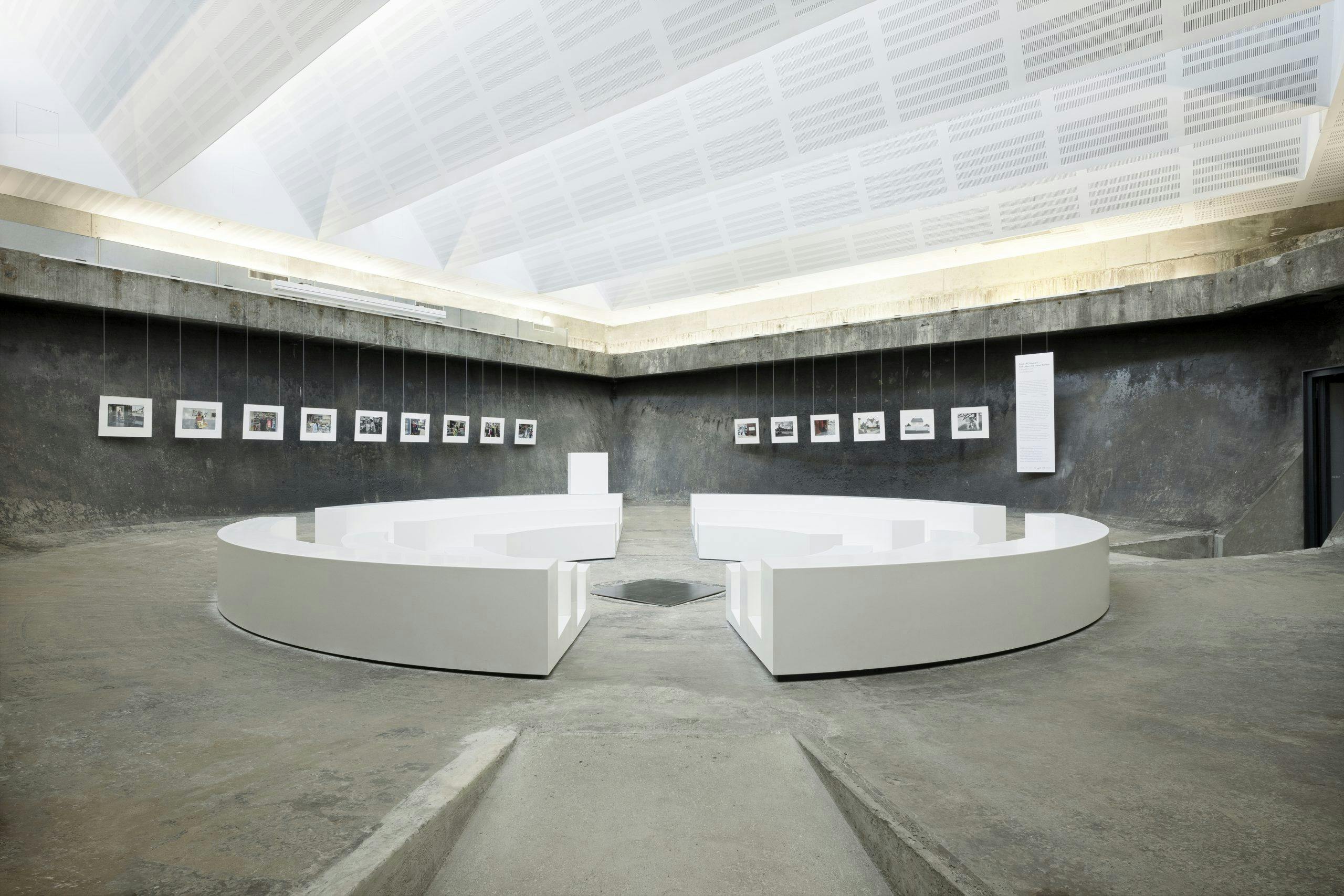 Ein heller, minimalistischer Ausstellungsraum mit rauen Betonwänden und einem geometrisch gestalteten, schräg verlaufenden Boden. In der Mitte stehen vier große, weiße, kreisförmig angeordnete Sitz- oder Präsentationseinheiten, die einen offenen Kreis bilden. An den Wänden hängen in gleichmäßigem Abstand kleine gerahmte Fotografien, die an dünnen Seilen von der Decke herabhängen. Der Raum wird von einer weißen, schräg verlaufenden Decke mit indirekter Beleuchtung erhellt. Die Atmosphäre ist ruhig, klar und modern.
