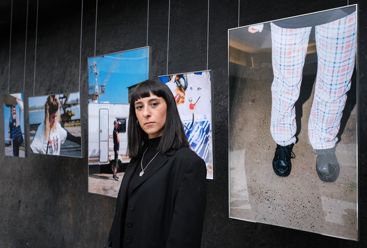 Amina Falah steht in einem Ausstellungsraum vor einer Reihe großformatiger Fotografien, die an Drähten von der Decke hängen. Sie trägt einen schwarzen Blazer, ein schwarzes Oberteil und eine silberne Halskette mit Anhänger. Ihr dunkles, glattes Haar ist kinnlang geschnitten mit geradem Pony. Die Bilder hinter ihr zeigen Szenen aus dem Alltag, darunter Porträts, Straßenszenen und ein Hundemotiv. Amina Falah blickt ernst und selbstbewusst in die Kamera, während die visuelle Gestaltung ihrer Ausstellung eine persönliche und dokumentarische Bildsprache erkennen lässt.