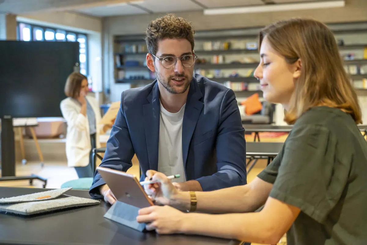 Ein junger Mann im blauen Sakko und eine junge Frau in einem dunkelgrünen Oberteil sitzen an einem Tisch in einem modernen, offenen Büro oder Coworking Space. Die Frau hält ein Tablet in der Hand, während der Mann aufmerksam zuhört und spricht. Im Hintergrund steht eine weitere Frau mit Smartphone am Ohr vor einem großen Monitor. Die Szene vermittelt konzentrierte Zusammenarbeit in kreativer Arbeitsumgebung.