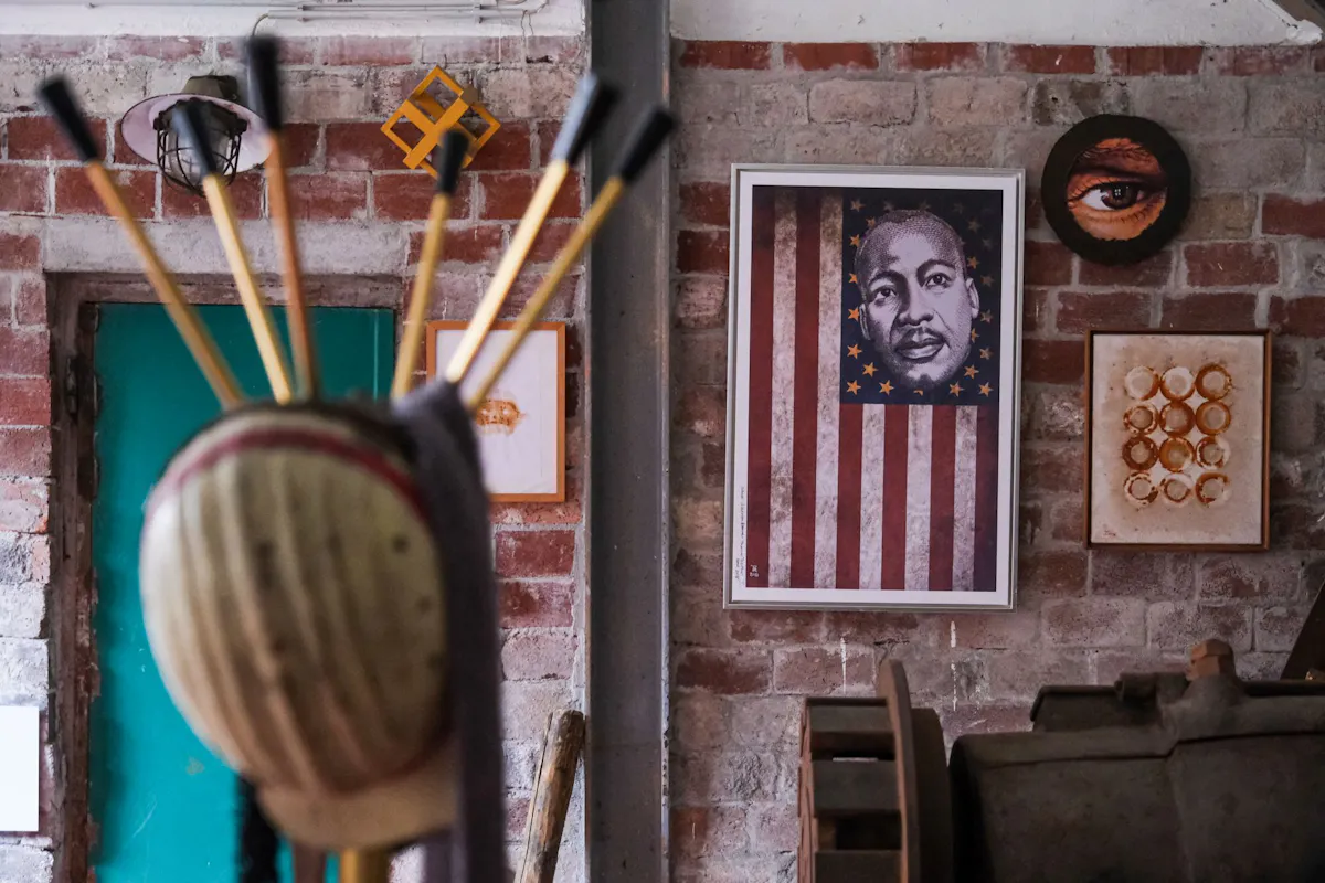 Blick in einen Raum mit Backsteinwänden, in dem mehrere Kunstwerke ausgestellt sind. Im rechten Bildbereich hängt ein gerahmtes Kunstwerk mit der Darstellung des Gesichts von Martin Luther King Jr., eingebettet in ein stilisiertes Motiv der US-Flagge. Daneben sind weitere Kunstwerke zu sehen: ein rundes Bild mit einem realistisch gemalten Auge, eine Arbeit mit ringförmigen Abdrücken auf Stoff und eine kleinere Grafik in Orange. Im Vordergrund steht eine skulpturale Holzfigur mit aufgesetzten Pfeilen, unscharf aufgenommen. Der Raum wirkt wie ein Atelier oder ein alternativer Ausstellungsort.