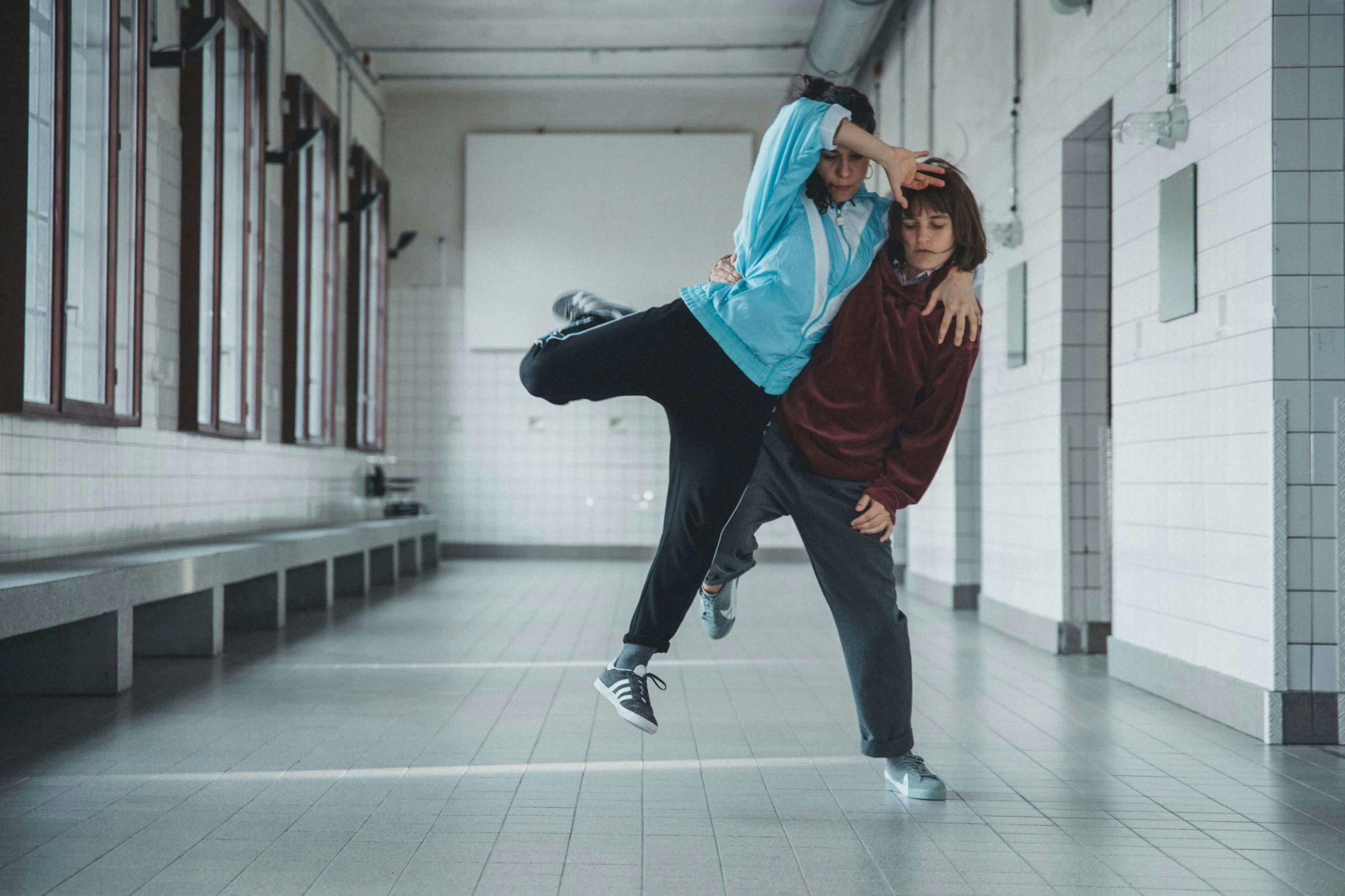 Zwei Tänzerinnen führen eine akrobatische Tanzpose in einem weiß gekachelten Raum mit industriellem Flair aus. Die linke Person trägt eine blaue Trainingsjacke und schwarze Hose, hebt ein Bein in die Luft und stützt sich mit einem Arm auf der Schulter der anderen ab. Die rechte Person trägt eine weinrote Jacke und graue Hose und trägt die Partnerin mit gebeugtem Knie. Der Raum wirkt wie eine ehemalige Umkleide mit großen Fenstern, grauen Fliesen, langen Sitzbänken und einer nüchternen Atmosphäre.