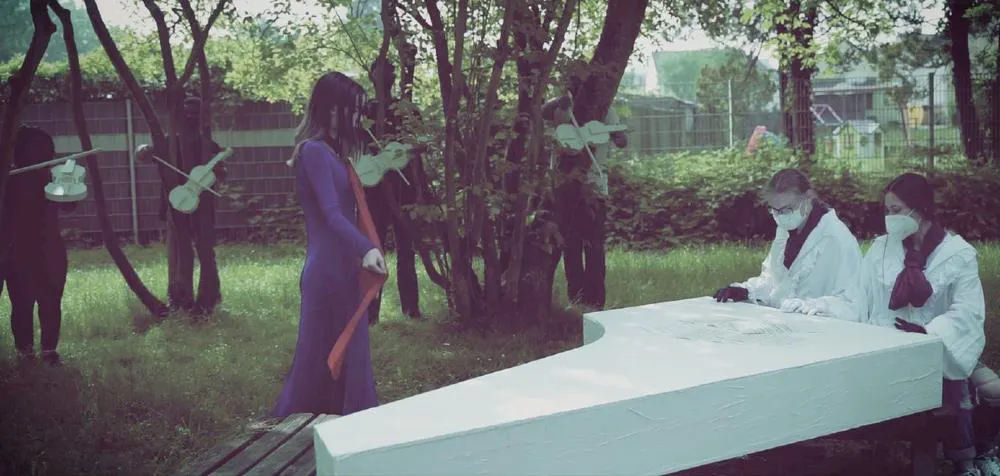 In einer inszenierten Szene im Freien stehen mehrere Personen in fantasievollen Kostümen. Im Vordergrund trägt eine Person ein violettes Kleid mit rotem Schal und steht vor einem weißen, klavierförmigen Objekt, an dem zwei andere Personen in weißen Kitteln und mit Mundschutz sitzen. Im Hintergrund stehen schwarz gekleidete Personen mit ausgeschnittenen Geigen-Silhouetten vor Bäumen. Die Atmosphäre wirkt geheimnisvoll und künstlerisch.
