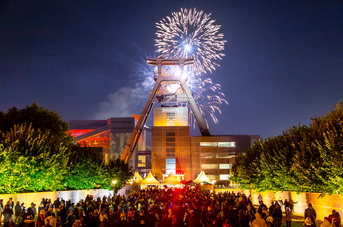 Bei Nacht versammeln sich viele Menschen vor dem beleuchteten Förderturm der Zeche Zollverein in Essen. Über dem markanten Industriedenkmal erleuchtet ein farbenprächtiges Feuerwerk den Himmel. Links und rechts des Weges stehen Bäume und Zelte, die stimmungsvoll angestrahlt sind. Die Szene wirkt festlich und lebendig.