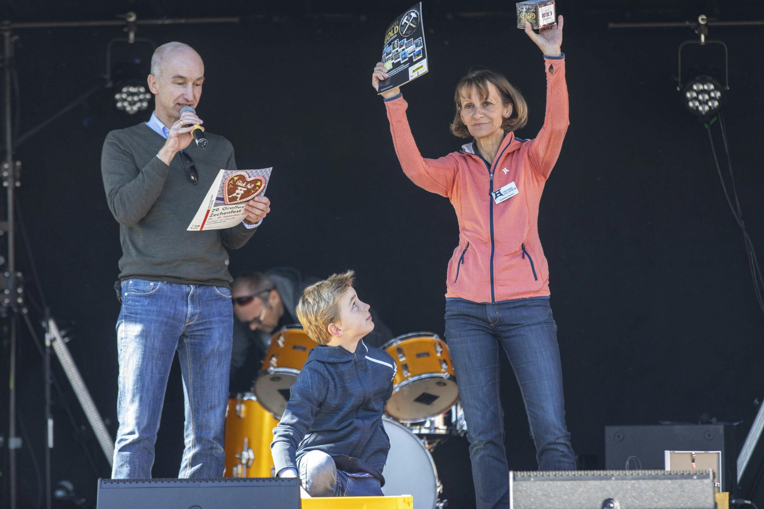 Auf einer Bühne stehen drei Personen vor einem schwarzen Hintergrund mit Schlagzeug im Hintergrund. Links spricht ein Mann in Jeans und dunklem Pullover in ein Mikrofon und hält ein Plakat mit der Aufschrift Zechenfest. Rechts daneben steht eine Frau in orangener Jacke und hebt stolz zwei Gewinnartikel in die Höhe, darunter ein Schild mit der Aufschrift Gold. Zwischen den beiden kniet ein Junge mit blonder Frisur in blauer Jacke und blickt zur Frau auf. Die Szene wirkt wie eine Preisverleihung oder Siegerehrung bei einer Veranstaltung.