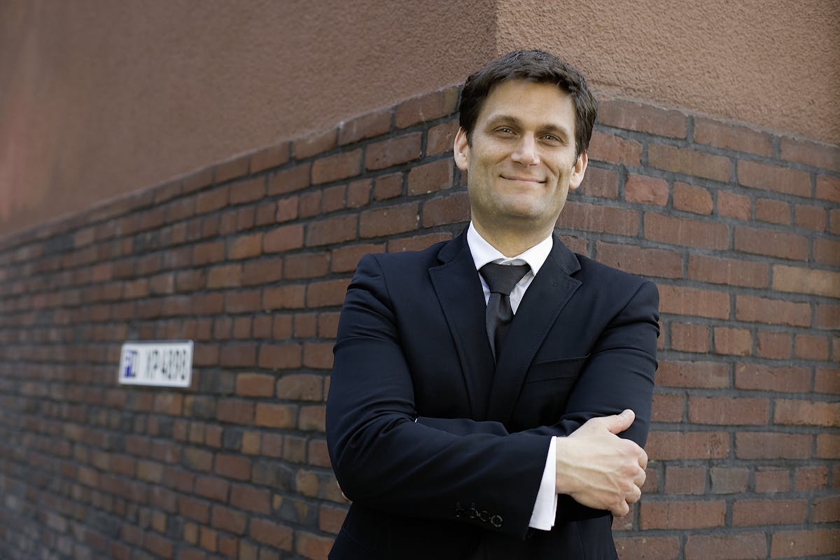 Ein Mann mit dunklem Haar trägt einen schwarzen Anzug mit weißem Hemd und schwarzer Krawatte. Er steht lächelnd und mit verschränkten Armen vor einer Ziegelmauer an einer Gebäudeecke. Im Hintergrund ist ein Schild mit der Aufschrift P1 WP-4808 zu sehen. Die Szene wirkt selbstbewusst und urban.