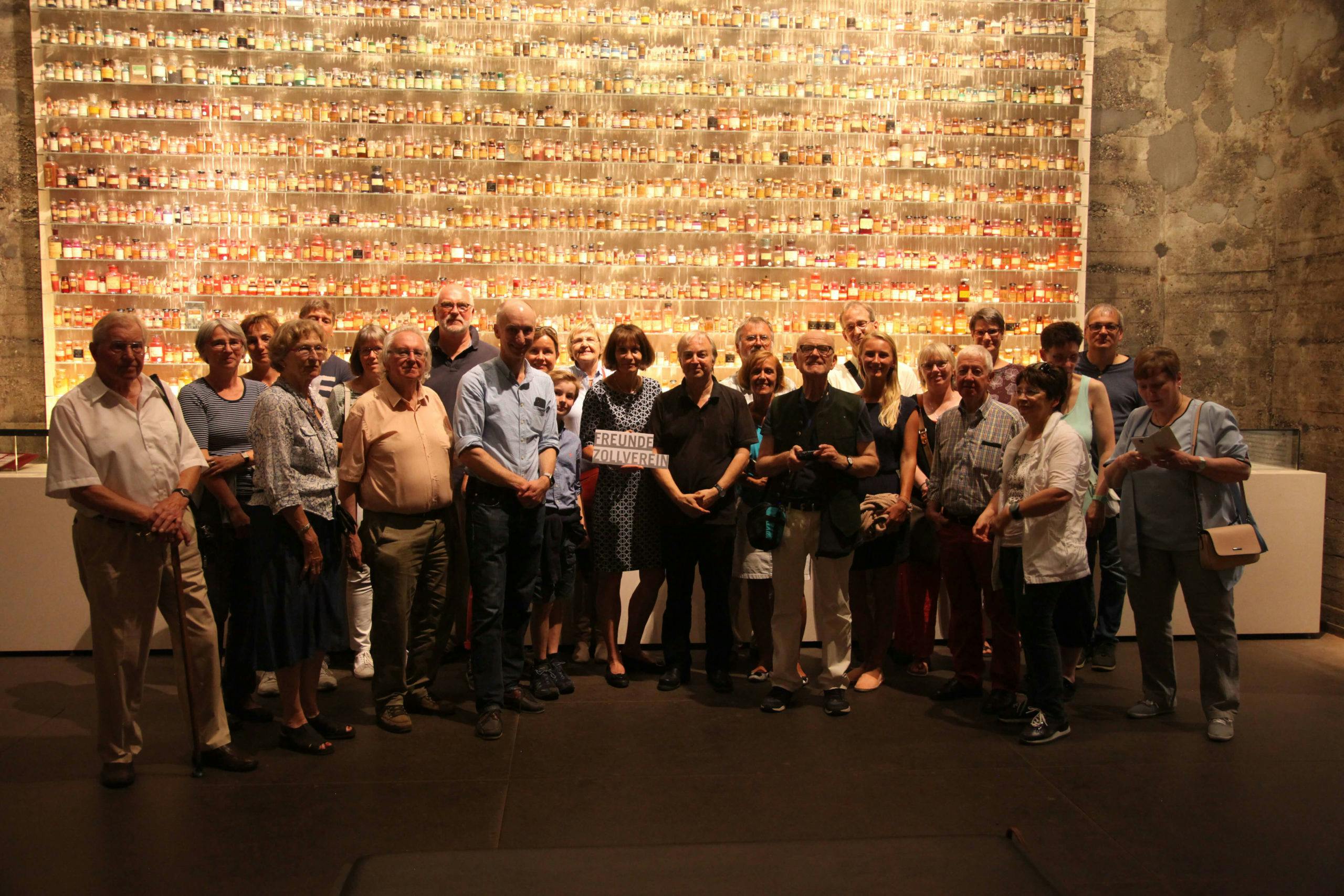 Eine Gruppe von etwa 25 Personen unterschiedlichen Alters steht dicht beieinander vor einer beleuchteten Wandinstallation, die aus hunderten kleiner Gläser in Reihen besteht, gefüllt mit verschiedenen Substanzen oder Materialien. Die Menschen lächeln in Richtung Kamera, einige haben die Arme verschränkt oder um andere gelegt. In der Mitte hält eine Frau ein Schild mit der Aufschrift Freunde Zollverein. Die Szene spielt sich in einem Ausstellungsraum mit industriell wirkender Betonwand im Hintergrund ab. Die Atmosphäre wirkt warm und gemeinschaftlich.
