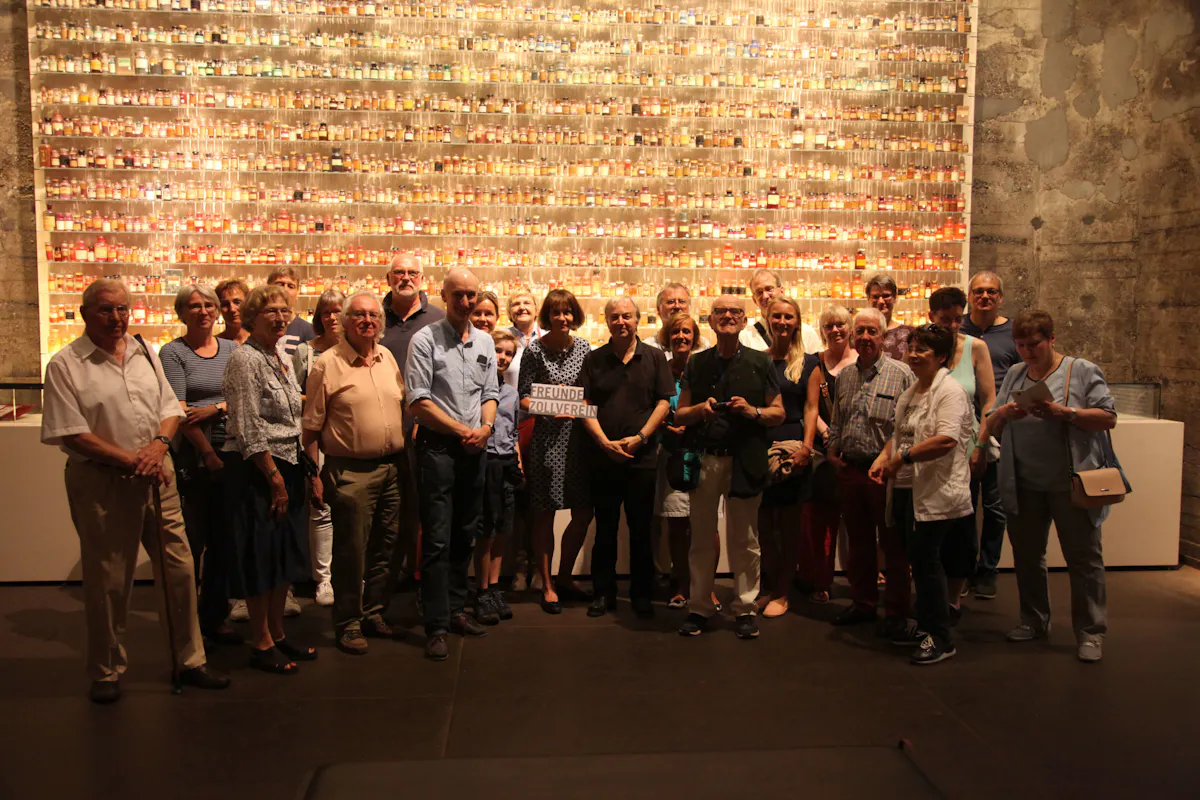 Eine Gruppe von etwa 25 Personen unterschiedlichen Alters steht dicht beieinander vor einer beleuchteten Wandinstallation, die aus hunderten kleiner Gläser in Reihen besteht, gefüllt mit verschiedenen Substanzen oder Materialien. Die Menschen lächeln in Richtung Kamera, einige haben die Arme verschränkt oder um andere gelegt. In der Mitte hält eine Frau ein Schild mit der Aufschrift Freunde Zollverein. Die Szene spielt sich in einem Ausstellungsraum mit industriell wirkender Betonwand im Hintergrund ab. Die Atmosphäre wirkt warm und gemeinschaftlich.