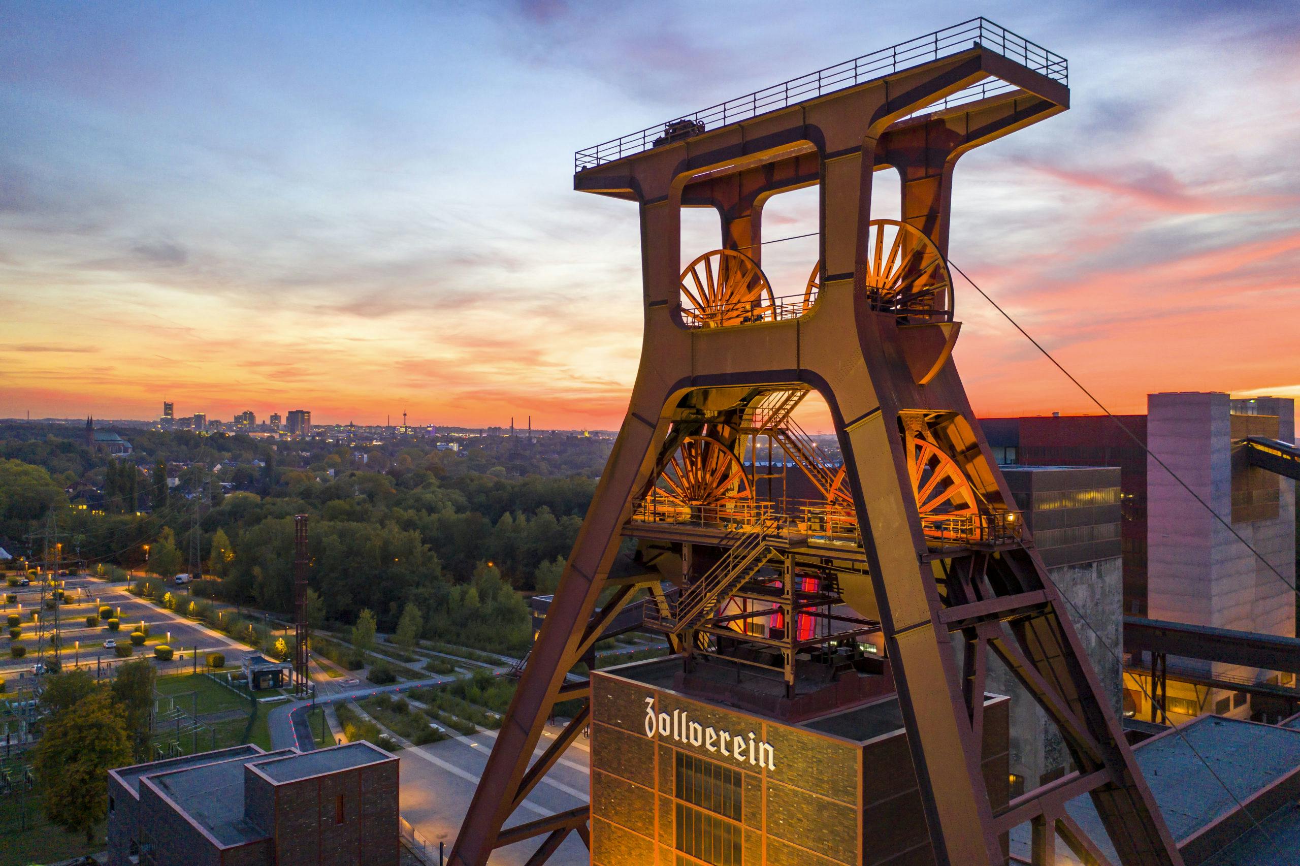 Blick auf den markanten Doppelbock-Förderturm der Zeche Zollverein in Essen bei Sonnenuntergang. Der imposante Turm aus Stahl mit seinen großen Seilscheiben ist zentral im Bild positioniert und wird von unten stimmungsvoll beleuchtet. Auf dem Gebäude unterhalb des Turms steht in weißer Schrift Zollverein. Im Hintergrund erstreckt sich ein weitläufiges Areal mit Bäumen, Wegen und historischen Industriegebäuden, dahinter die Silhouette der Stadt Essen. Der Himmel ist in kräftigen Farben von orange bis violett gefärbt und verleiht der Szene eine eindrucksvolle Atmosphäre.