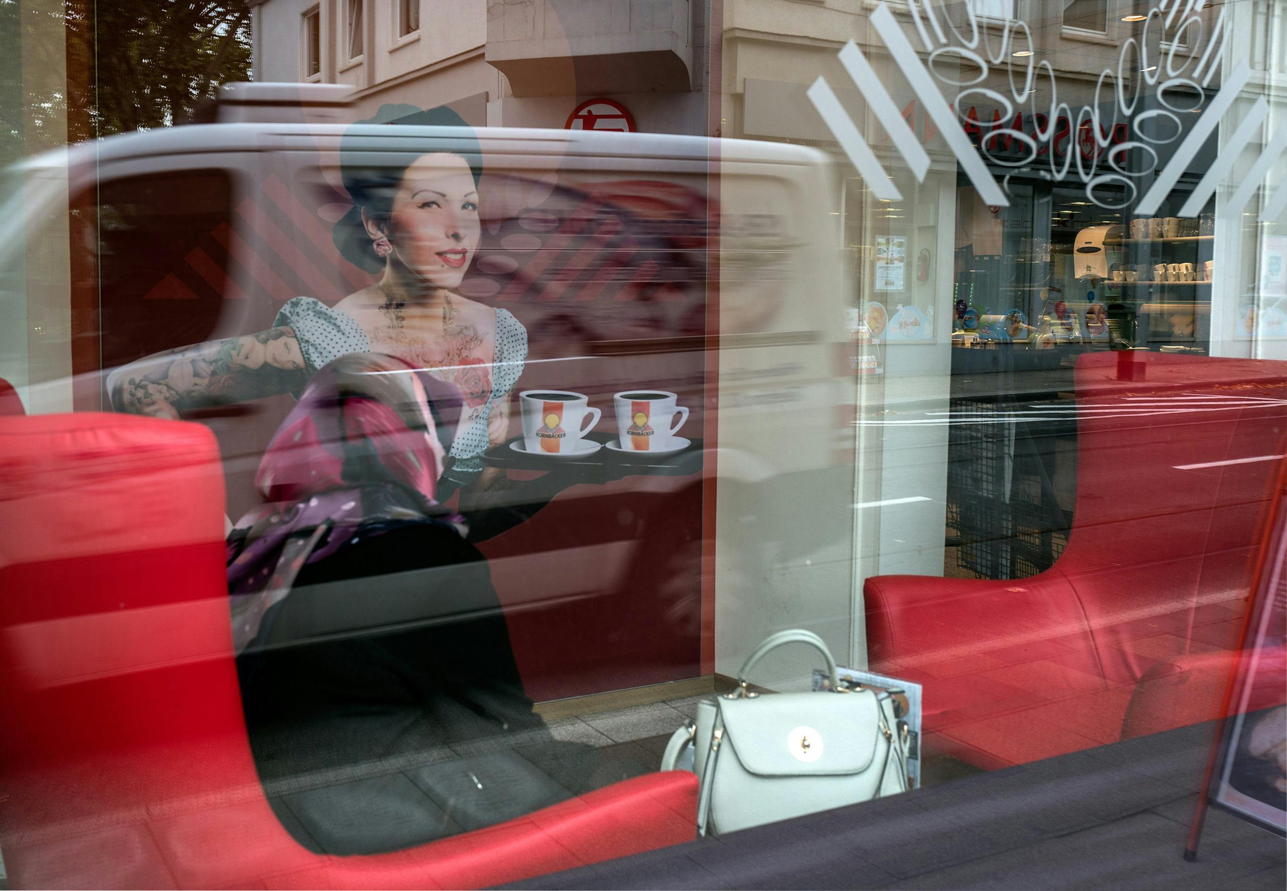 Blick durch eine Schaufensterscheibe auf die Inneneinrichtung eines Cafés mit roten Sitzmöbeln und einer hellen Handtasche. Auf der Scheibe spiegelt sich ein vorbeifahrender weißer Lieferwagen, in dem ein großflächiges Werbebild einer tätowierten Frau mit Tablett und zwei Kaffeetassen zu sehen ist. Auch Straßenszene und Gebäudefassaden spiegeln sich im Glas. Die Aufnahme wirkt durch die Spiegelungen vielschichtig und dynamisch.