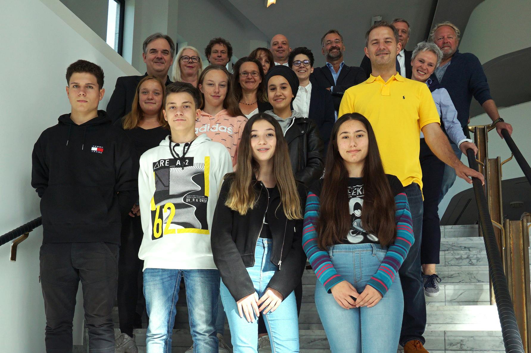 Eine Gruppe von Jugendlichen und Erwachsenen steht auf einer Marmortreppe in einem modernen Gebäude. Die Jugendlichen befinden sich in der vorderen Reihe, während die Erwachsenen in mehreren Reihen dahinter angeordnet sind. Alle blicken in die Kamera und lächeln oder schauen neutral. Die Szene wirkt wie ein Gruppenfoto im Rahmen eines Schul- oder Bildungsprojekts.