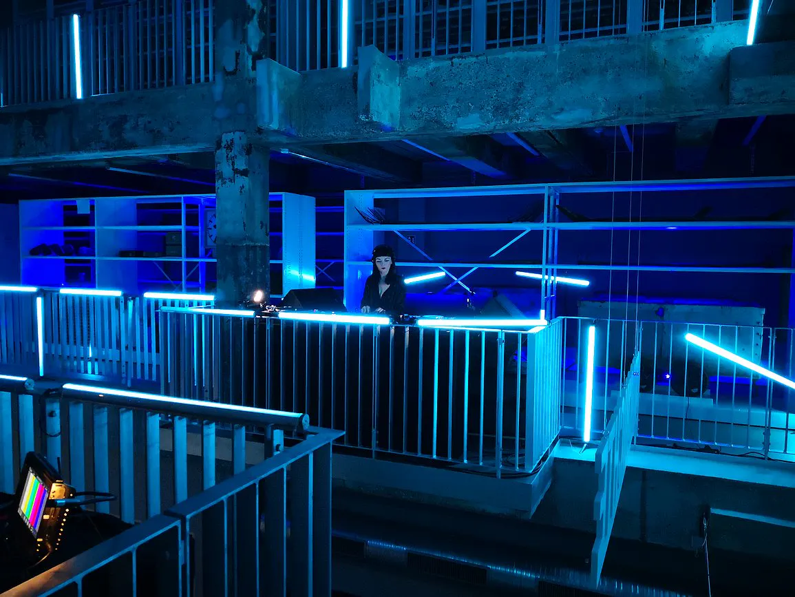 Eine Person steht an einem DJ-Pult in einem industriellen Gebäude mit mehreren Ebenen und Geländern. Die gesamte Szene ist in blaues Neonlicht getaucht, mit zahlreichen leuchtenden Lichtstäben, die futuristisch wirken. Die Atmosphäre erinnert an ein Club- oder Kunstevent in urbaner Umgebung.
