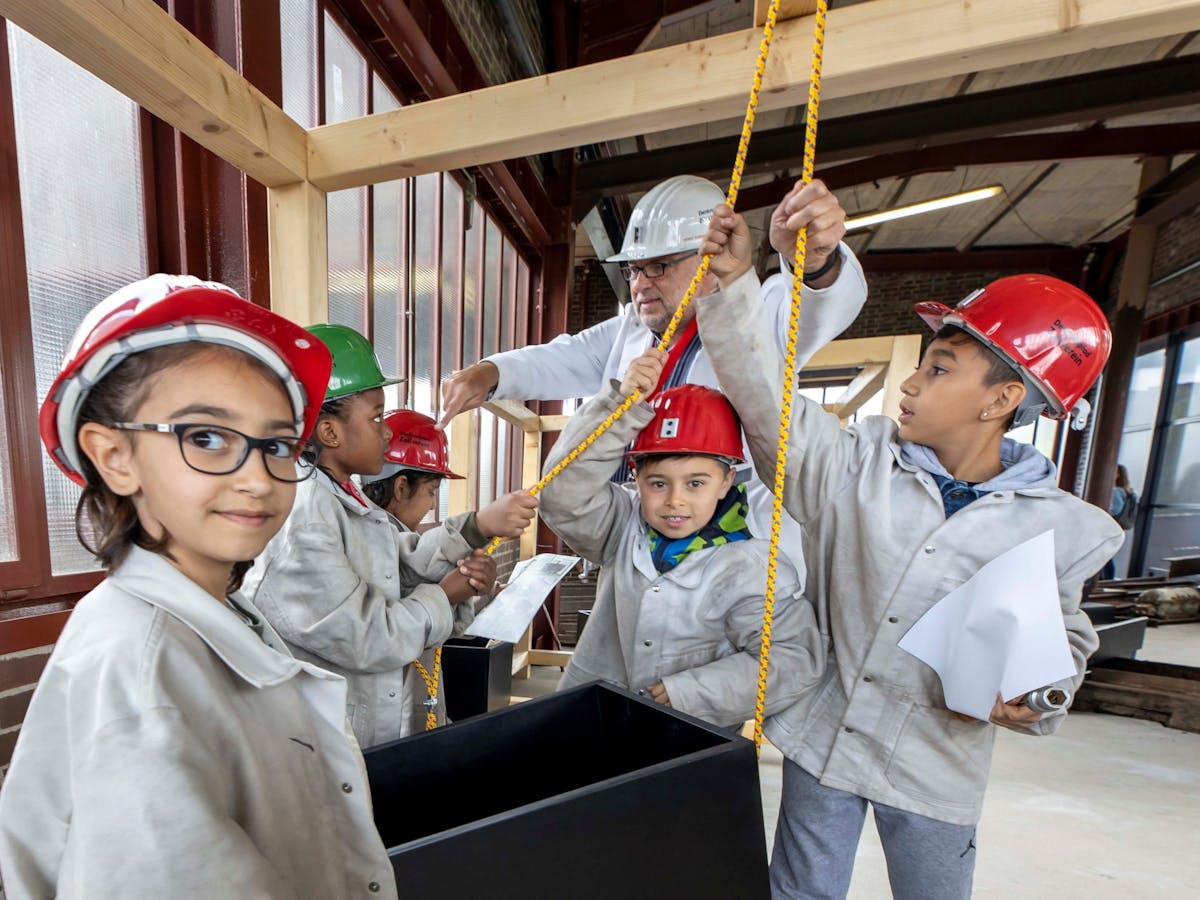 Eine Gruppe von Kindern mit bunten Schutzhelmen und grauen Arbeitsjacken nimmt an einer handwerklichen Mitmachaktion in einem überdachten, lichtdurchfluteten Raum teil. Ein älterer Mann mit weißem Helm und Schutzbrille erklärt ihnen den Umgang mit einem gelben Seilzug. Die Kinder wirken konzentriert und engagiert, einige halten Werkzeuge oder Papier in den Händen. Im Vordergrund blickt ein Mädchen mit Brille freundlich in die Kamera. Die Szene vermittelt einen pädagogischen Kontext mit handlungsorientiertem Lernen.