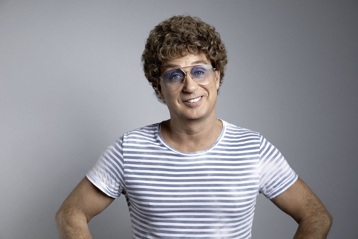 Ein lächelnder Mensch mit heller Haut und brauner, lockiger Perücke steht vor einem neutral grauen Hintergrund. Die Person trägt eine große, hellblaue Retro-Brille und ein kurzärmeliges T-Shirt mit weißen und grauen Querstreifen. Die Hände sind locker in die Hüften gestützt, der Gesichtsausdruck ist freundlich und selbstbewusst.