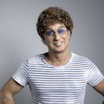 Ein lächelnder Mensch mit heller Haut und brauner, lockiger Perücke steht vor einem neutral grauen Hintergrund. Die Person trägt eine große, hellblaue Retro-Brille und ein kurzärmeliges T-Shirt mit weißen und grauen Querstreifen. Die Hände sind locker in die Hüften gestützt, der Gesichtsausdruck ist freundlich und selbstbewusst.