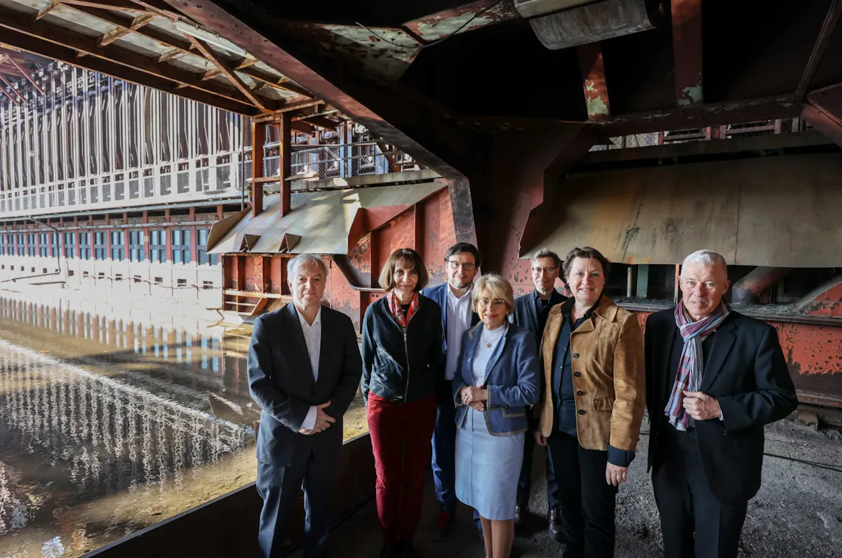 Gruppenfoto mit sieben Frauen und Männern in formeller Kleidung, die nebeneinander in einer historischen Industrieanlage der Kokerei Zollverein stehen. Im Hintergrund ist die beeindruckende Anlage mit rostroten Stahlkonstruktionen, Rohrleitungen und einer spiegelnden Wasserfläche zu sehen. Die Personen stehen unter einem weit auskragenden Metalldach und lächeln in Richtung Kamera. Die Szene wirkt wie ein offizieller Fototermin im Rahmen einer Führung oder Veranstaltung im Industriedenkmal.