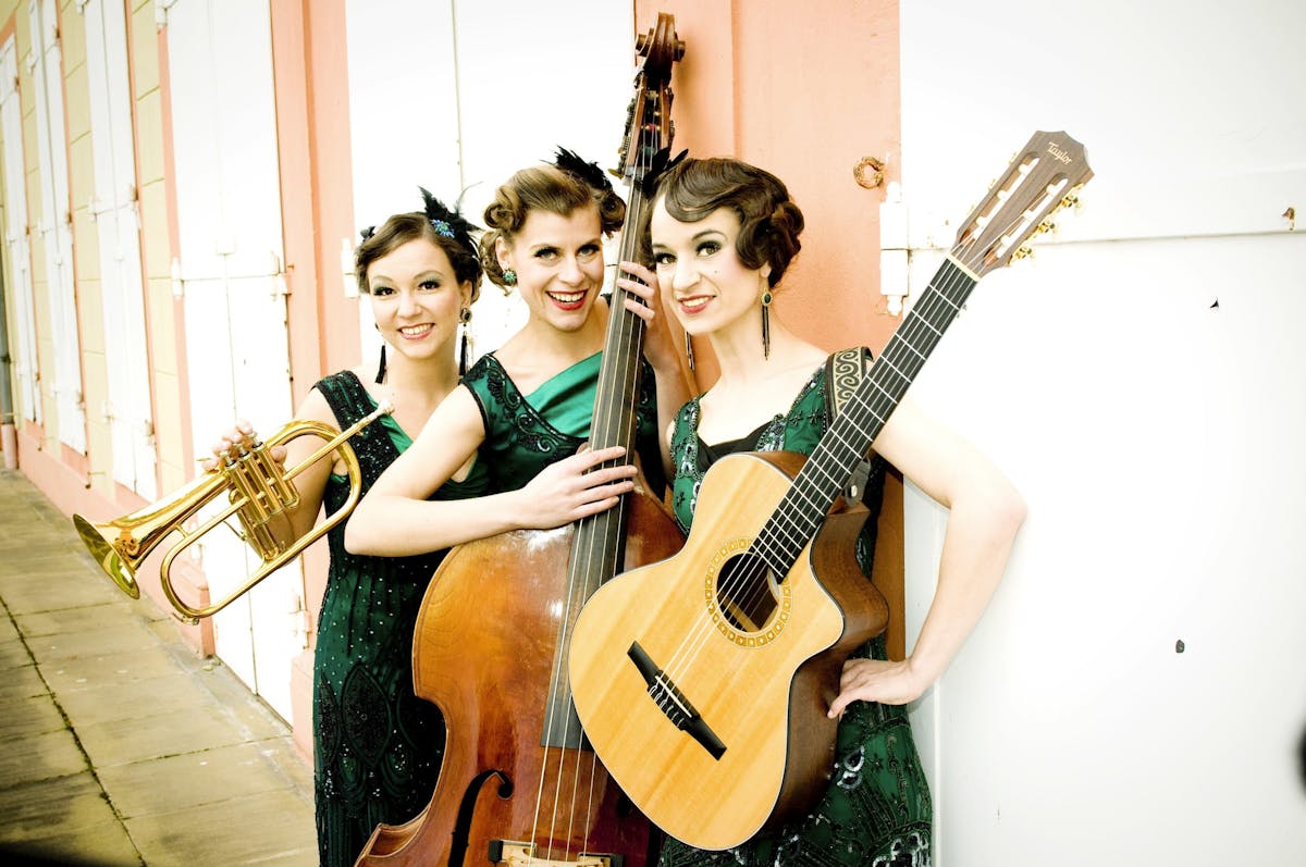 Drei junge Frauen in glitzernden grünen Kleidern stehen nebeneinander und lächeln in die Kamera. Jede von ihnen hält ein Musikinstrument: eine Trompete, einen Kontrabass und eine Gitarre. Sie haben aufwendige Vintage-Frisuren mit Haaraccessoires und tragen auffälligen Schmuck. Der Hintergrund zeigt eine helle Fassade mit orangefarbenen Säulen und geschlossenen weißen Fensterläden.