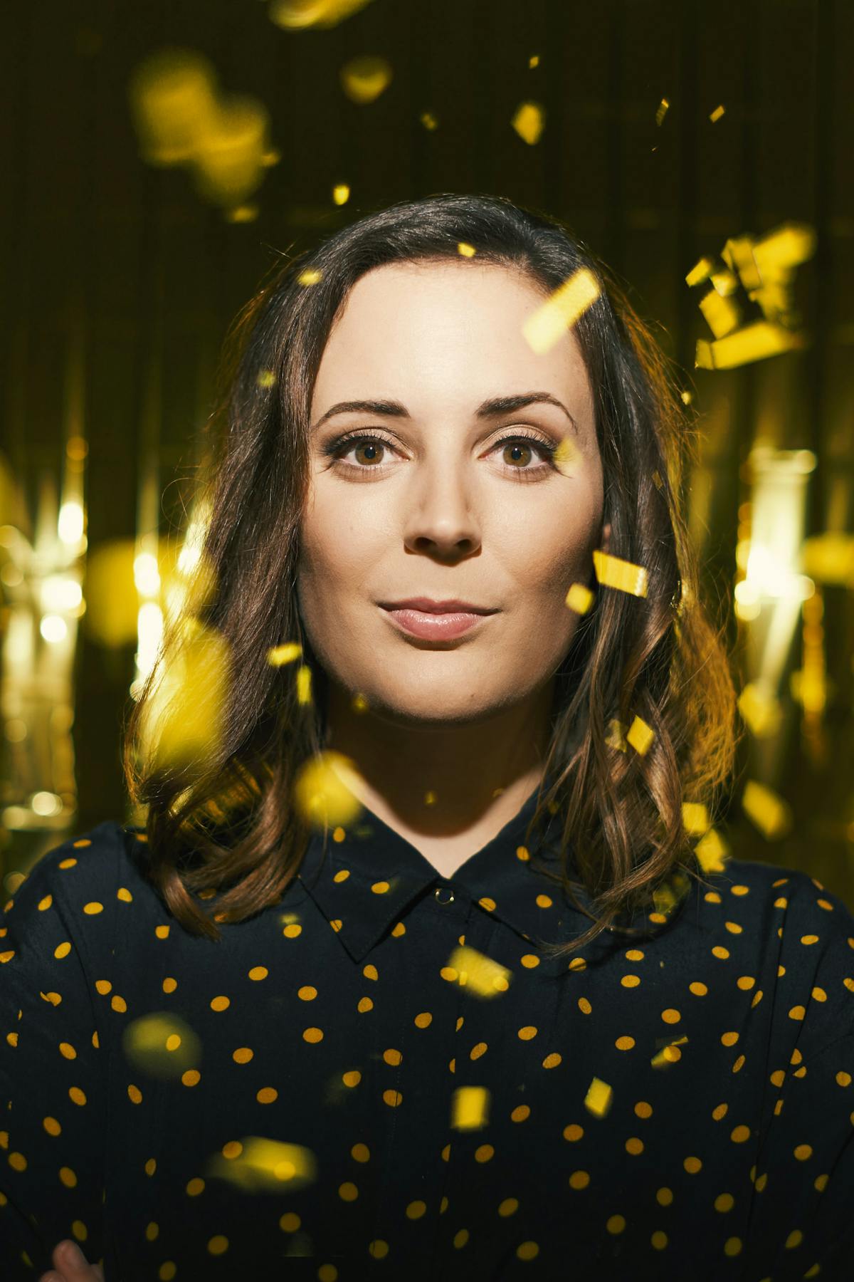 Eine Frau mit schulterlangen braunen Haaren und dunklem Hemd mit gelben Punkten blickt ruhig und direkt in die Kamera. Um sie herum fliegen goldene Konfetti-Streifen, die von oben herabfallen. Der Hintergrund ist dunkel und glänzend, was eine festliche und dramatische Stimmung erzeugt.