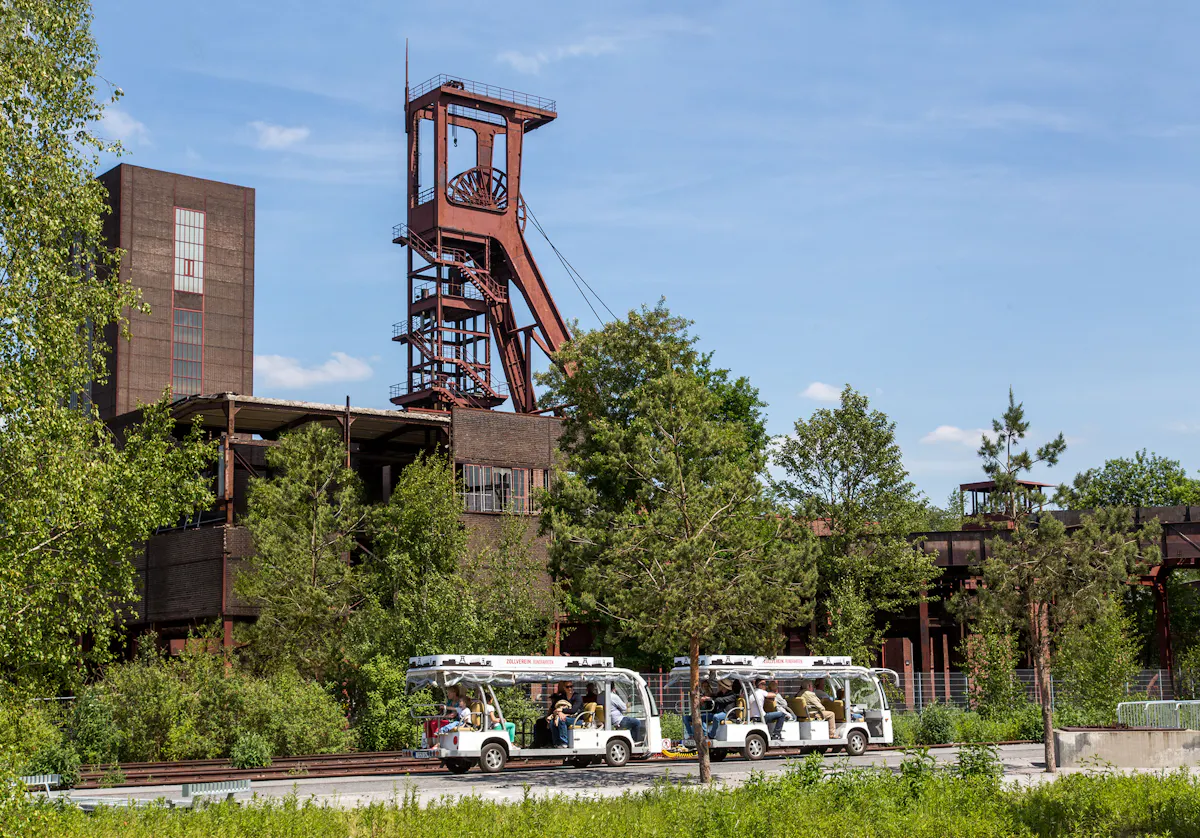 Zwei weiße Besucherfahrzeuge mit offenen Seiten und mehreren Sitzreihen fahren bei sonnigem Wetter durch eine grüne Parklandschaft. Im Hintergrund steht ein historisches Industriegebäude mit einem markanten roten Förderturm, teilweise von Bäumen umgeben. Das Gebäude gehört zum UNESCO-Welterbe Zollverein in Essen und symbolisiert Industriekultur. Der Himmel ist blau mit wenigen Wolken, was die Szene freundlich und einladend wirken lässt.
