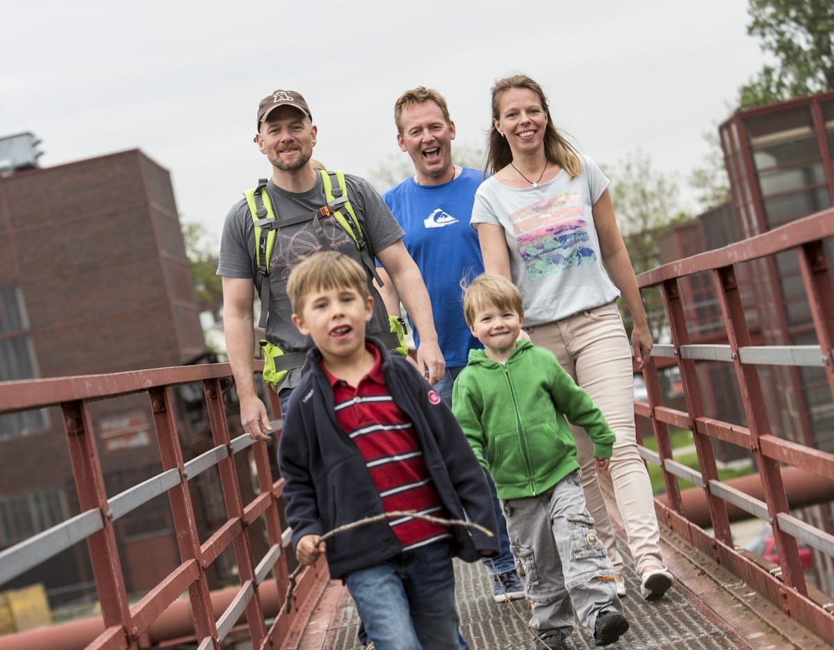 Eine fröhliche Gruppe aus zwei Erwachsenen, einer Frau und zwei Männern, sowie drei Kindern geht gemeinsam über eine rote Metallbrücke. Die Kinder laufen vorneweg, zwei Jungen im Grundschulalter lachen in die Kamera, einer trägt einen Stock in der Hand. Dahinter folgen die Erwachsenen in legerer Freizeitkleidung, teilweise mit Rucksäcken. Im Hintergrund sind rote Industriebauten und grüne Bäume zu erkennen, was auf ein ehemaliges Industriegelände hinweist. Die Stimmung ist lebhaft und familiär.