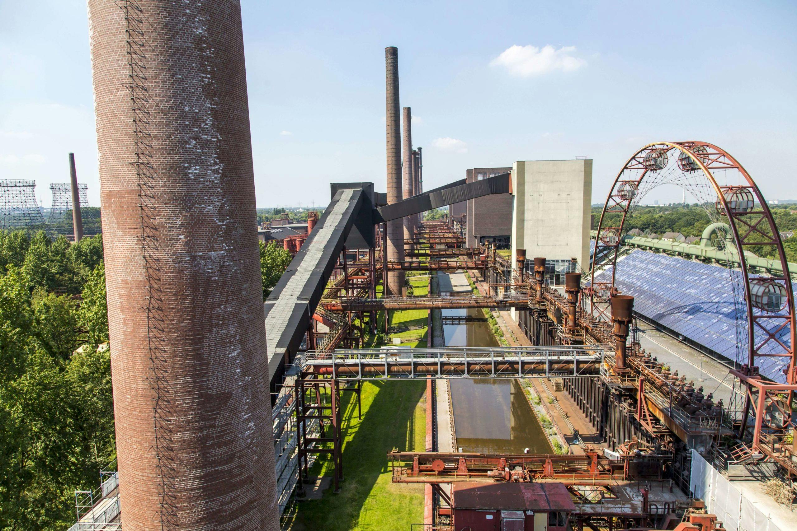 Luftaufnahme des ehemaligen Kokereigeländes Zollverein bei Sonnenschein. Im Vordergrund ragt ein hoher, runder Schornstein aus Backstein empor, daneben verlaufen rostige Rohrleitungen und Förderanlagen entlang mehrerer Wasserbecken. Im rechten Bildbereich steht ein großes, halbtransparentes Solardach und dahinter ein auffälliges Riesenrad aus Stahl mit verglasten Gondeln. Im Hintergrund sind weitere Industriegebäude, Schlote und grüne Baumflächen zu sehen. Die Szene zeigt den Kontrast zwischen Industriearchitektur und moderner Umnutzung.