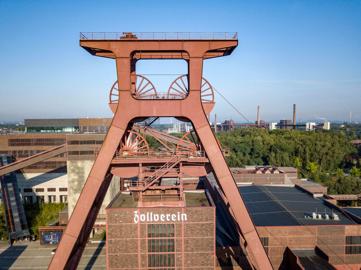 Luftaufnahme des markanten Förderturms der Zeche Zollverein bei klarem, sonnigem Wetter. Der rostrote Turm mit seinen großen Seilscheiben steht im Zentrum des Bildes und prägt das historische Industriegelände. Darunter ist ein Backsteingebäude mit dem Schriftzug Zollverein zu sehen. Im Hintergrund erstreckt sich eine grüne Baumlandschaft sowie weitere Industrieanlagen und Schornsteine, die bis zum Horizont reichen. Die Szene vermittelt einen Eindruck von industrieller Geschichte und landschaftlicher Weite.