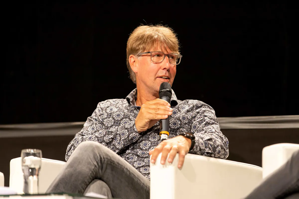 Ein Mann mittleren Alters mit blond-braunen Haaren und Brille sitzt in einem weißen Sessel auf einer Bühne. Er trägt ein gemustertes Hemd und graue Jeans und hält ein Mikrofon in der rechten Hand, während er zur Seite blickt und spricht. Auf dem Tisch vor ihm stehen ein Glas Wasser und ein Notizblock. Der Hintergrund ist dunkel, wodurch der Mann und die Szene im Vordergrund stark hervorgehoben werden.