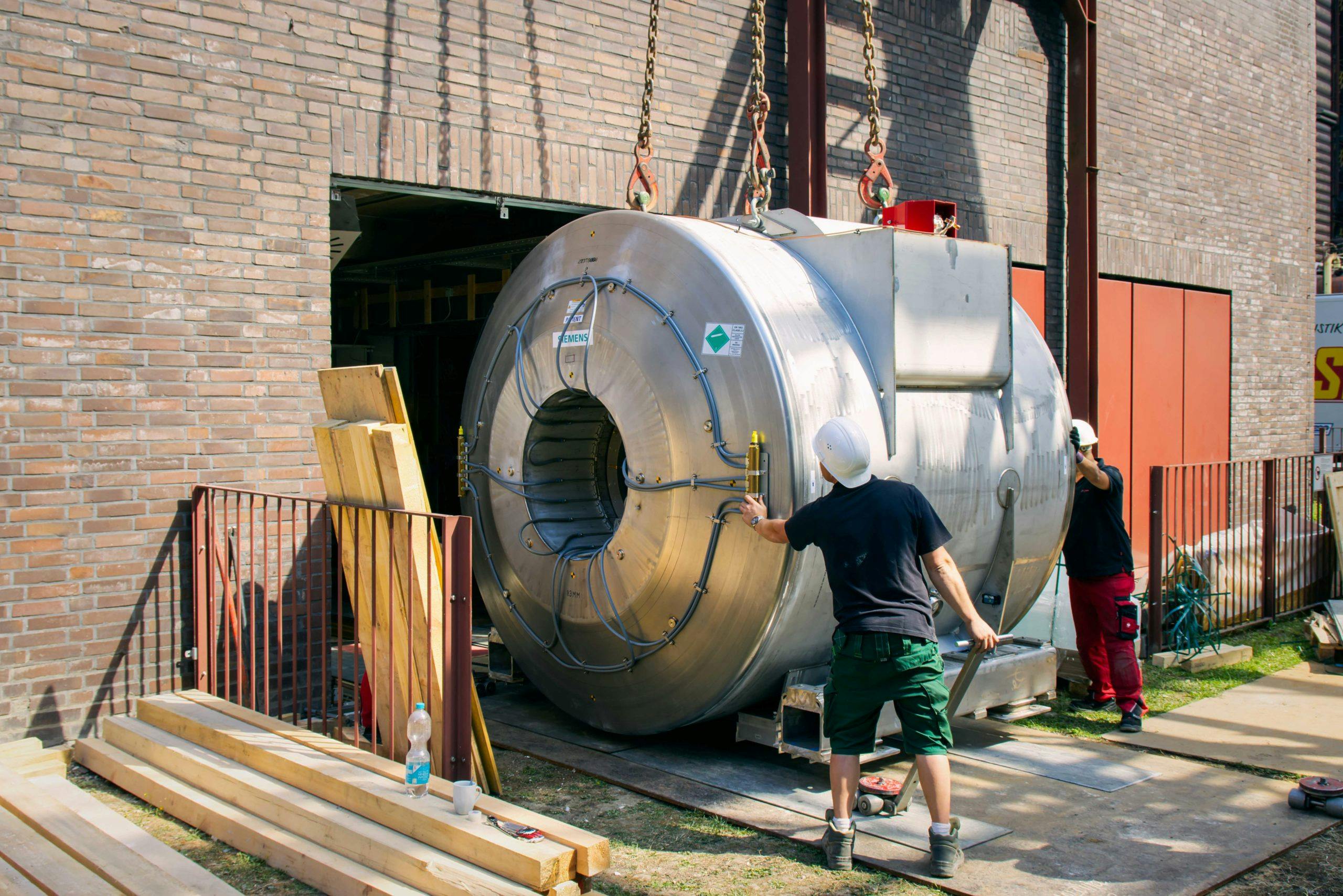 Zwei Männer mit Schutzhelmen und Arbeitskleidung manövrieren ein großes, zylinderförmiges Metallgerät mithilfe eines Krans durch eine geöffnete Tür eines Backsteingebäudes. Das Gerät hat eine zentrale Öffnung und ist mit Leitungen und Anschlüssen versehen. Es trägt das Siemens-Logo und ein Gefahrensymbol. Vor dem Gebäude liegen Holzbalken und Bauutensilien, darunter eine Wasserflasche und eine Tasse. Die Szene spielt bei sonnigem Wetter und vermittelt eine aktive Baustellenatmosphäre.