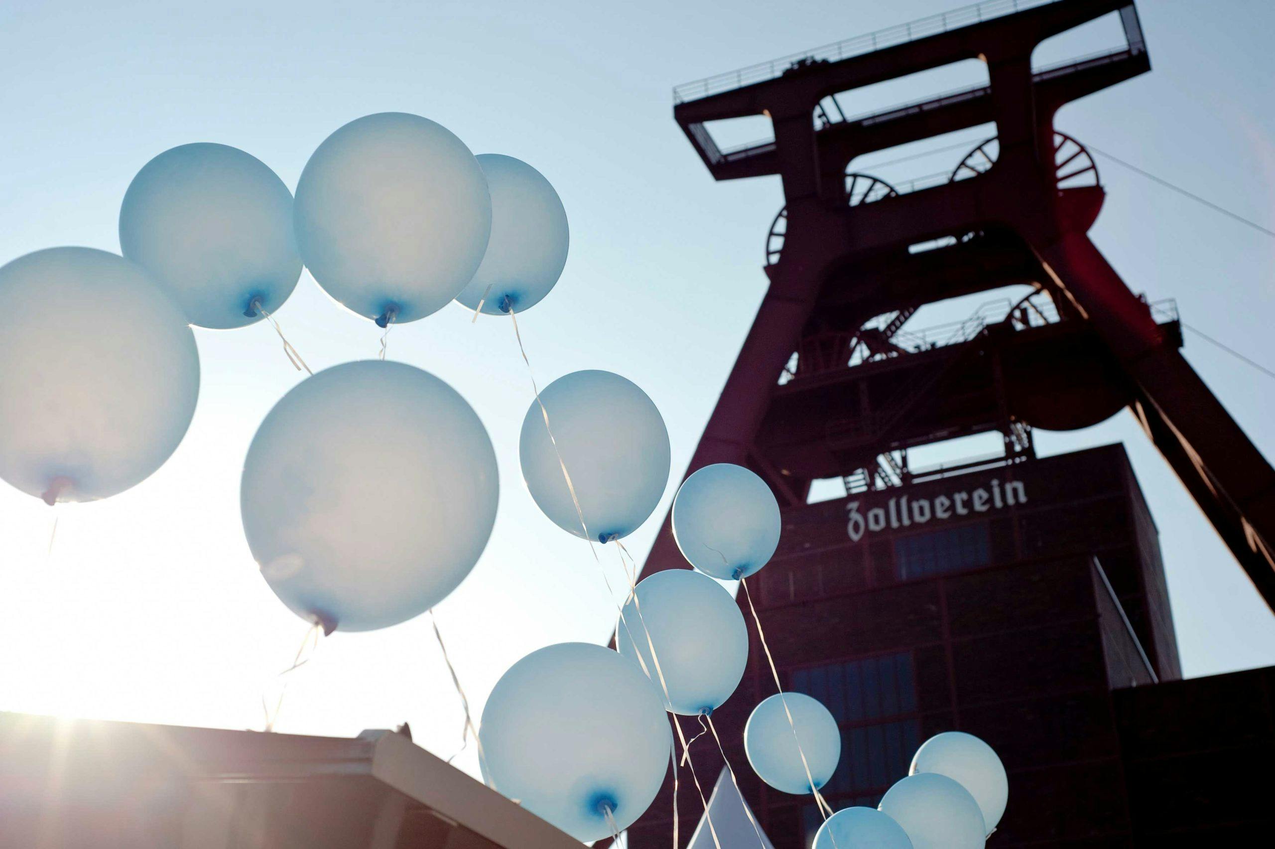 Mehrere hellblaue Luftballons steigen in den Himmel vor der Kulisse des ikonischen Fördergerüsts der Zeche Zollverein in Essen. Die Ballons sind an weißen Schnüren befestigt und werden von unten links ins Bild geführt. Im Hintergrund ist die rote Stahlkonstruktion des Förderturms mit der Aufschrift Zollverein zu sehen. Die Sonne scheint von links unten ins Bild und erzeugt eine helle Lichtstimmung mit weichen Schatten. Der Himmel ist klar und blau.