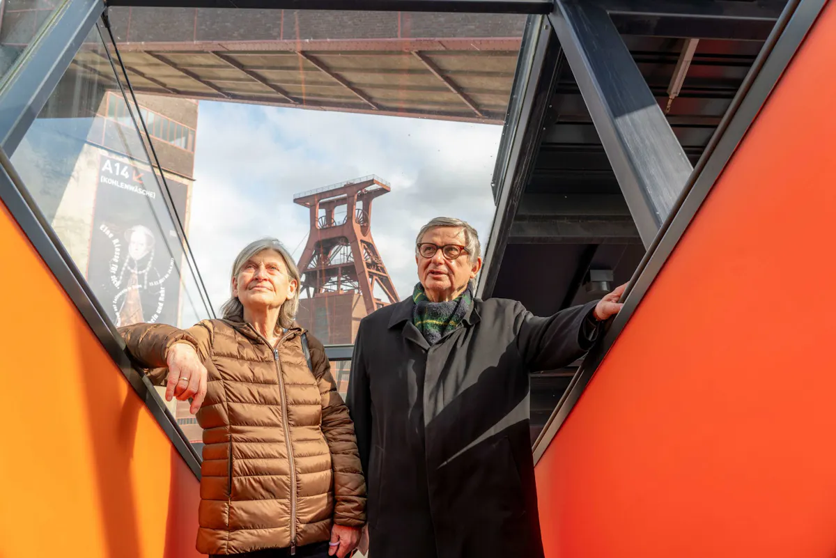 Ein älteres Paar steht auf einer modernen, orangefarbenen Rolltreppe des Ruhr Museums auf dem Gelände der Zeche Zollverein in Essen. Beide blicken nach oben, die Frau trägt eine braune Steppjacke, der Mann einen dunklen Mantel mit Schal und Brille. Im Hintergrund ist das markante rote Fördergerüst der Zeche sowie ein Plakat mit der Aufschrift A14 Kohlenwäsche zu sehen. Die Szene ist bei Tageslicht aufgenommen, der Himmel ist leicht bewölkt.