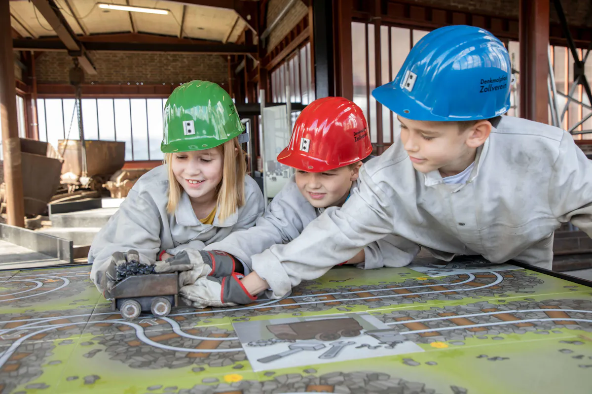 Drei Kinder in grauen Schutzjacken und bunten Helmen – grün, rot und blau – spielen gemeinsam an einem großen, farbigen Tisch mit einem grafisch gestalteten Bergwerksplan. Sie bewegen eine kleine Kohlelore mit schwarzer Füllung über aufgemalte Gleise. Alle wirken konzentriert und fröhlich. Die Szene spielt in einer Halle mit industrieller Architektur und großen Fenstern. Im Hintergrund stehen echte Grubenhunte. Die Umgebung vermittelt eine Mischung aus spielerischem Lernen und authentischer Bergbauatmosphäre auf dem Denkmalpfad Zollverein.