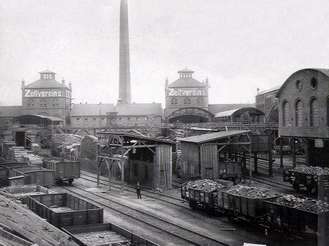 Historische Schwarz-Weiß-Aufnahme des Zechengeländes Zollverein mit Blick auf die Schächte Zollverein I und II. Die zentralen Gebäude tragen deutlich sichtbare Beschriftungen mit Zollverein I und Zollverein II. Im Vordergrund befinden sich Gleise mit offenen Güterwaggons, die mit Kohle beladen sind. Dazwischen stehen einfache Holzunterstände und Förderanlagen. In der Mitte des Bildes ragt ein hoher Schornstein auf, dahinter sind langgestreckte Backsteinbauten zu sehen. Eine einzelne Person steht auf den Gleisen, was die Dimensionen der Anlage verdeutlicht. Die Szene zeigt das geschäftige industrielle Umfeld einer aktiven Zeche um die Jahrhundertwende.
