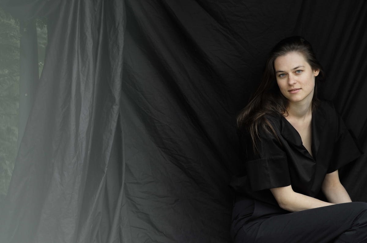 Eine junge Frau mit langen braunen Haaren sitzt vor einem schwarzen, leicht gerafften Stoffhintergrund. Sie trägt ein locker geschnittenes schwarzes Oberteil und schwarze Hose. Ihr Gesicht ist leicht zur Seite geneigt, der Blick ist ruhig und direkt zur Kamera gerichtet. Die Lichtstimmung ist weich und natürlich, was dem Porträt eine ruhige und nachdenkliche Atmosphäre verleiht.