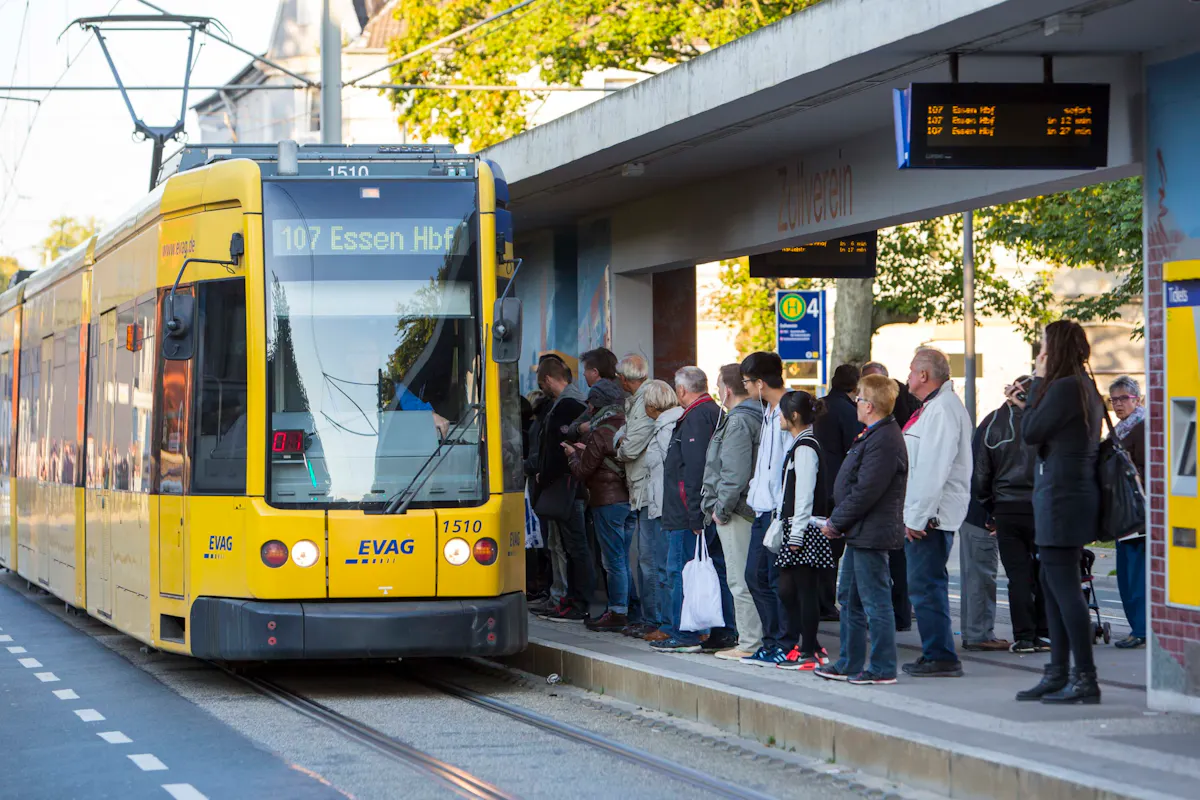 An einer Straßenbahnhaltestelle mit dem Namen Zollverein steht eine gelbe Straßenbahn der Linie 107 mit Ziel Essen Hauptbahnhof. Zahlreiche Fahrgäste warten auf dem Bahnsteig in einer langen Reihe, viele in herbstlicher Kleidung. Einige Menschen steigen gerade ein, während andere geduldig anstehen. Über dem Bahnsteig hängt eine digitale Anzeigetafel mit Abfahrtszeiten, im Hintergrund sind Bäume und Wohnhäuser zu sehen. Rechts im Bild befindet sich ein Fahrkartenautomat. Die Szene vermittelt Alltag und Mobilität im städtischen Nahverkehr.