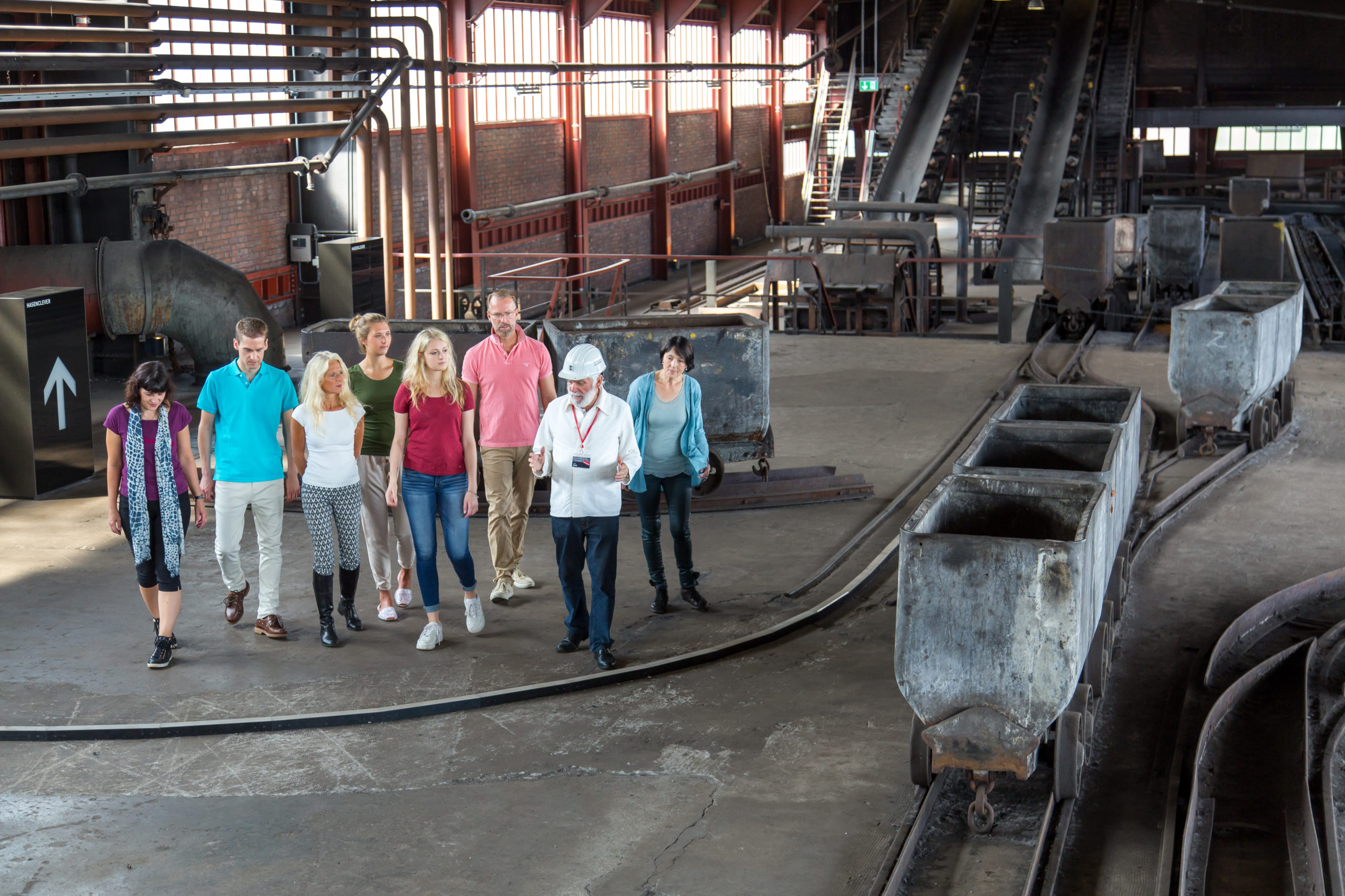 Eine Besuchergruppe bestehend aus neun Personen wird von einem Mann mit weißem Helm und Namensschild durch eine historische Industriehalle geführt. Die Gruppe geht an alten Förderwagen auf Schienen vorbei, die in einem großen, lichtdurchfluteten Raum mit industrieller Ausstattung und hohen Fenstern stehen. Die Szenerie vermittelt den Eindruck einer geführten Tour durch ein ehemaliges Bergwerk.