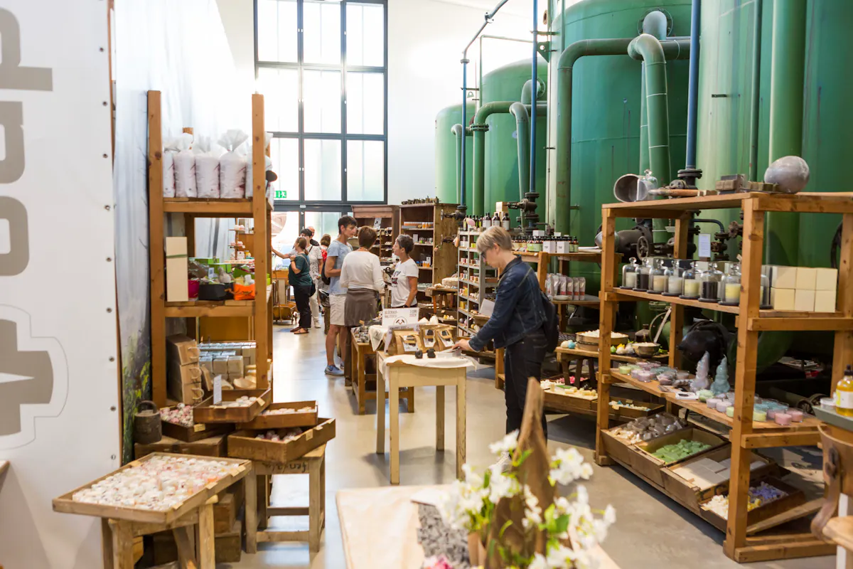 Innenaufnahme eines Verkaufsraums mit industriellem Flair, in dem handgefertigte Produkte wie Seifen, Kerzen und Kosmetikartikel auf rustikalen Holztischen und Regalen ausgestellt sind. Im Hintergrund befinden sich große grüne Tanks und Rohrleitungen, die an eine ehemalige Fabrik erinnern. Mehrere Menschen, überwiegend Frauen, schlendern durch die Gänge, betrachten die Ware oder unterhalten sich. Das Licht fällt durch hohe Fenster und schafft eine freundliche, offene Atmosphäre. Die Mischung aus Naturprodukten und industrieller Kulisse vermittelt einen Eindruck von nachhaltigem Design und regionaler Handwerkskunst.