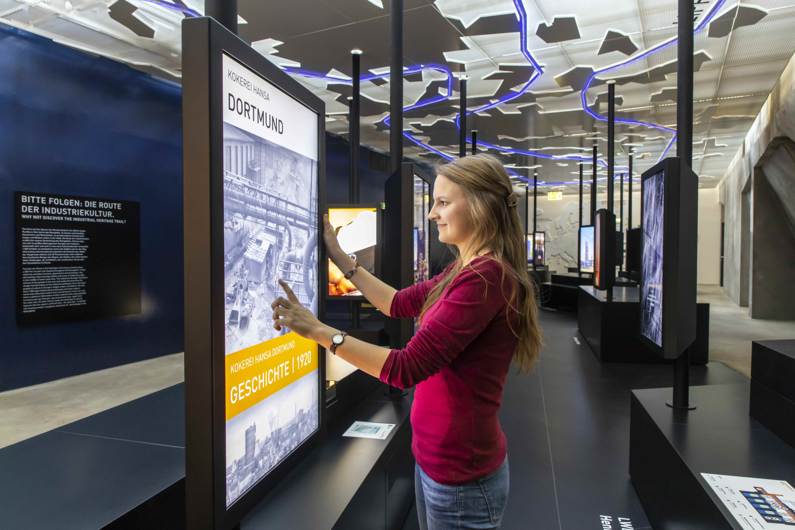 Eine junge Frau mit langen, hellbraunen Haaren steht in einer modernen Ausstellungshalle vor einem großen interaktiven Bildschirm. Sie trägt ein weinrotes Oberteil und berührt den Touchscreen mit beiden Händen. Auf dem Bildschirm ist eine historische Fotografie mit dem Titel Kokerei Hansa Dortmund und der Jahreszahl 1920 zu sehen. Im Hintergrund sind weitere interaktive Stationen und ein stilisiertes Deckenlichtmuster in Blau zu erkennen. Links an der Wand befindet sich ein erklärender Text mit dem Titel Bitte folgen: Die Route der Industriekultur.