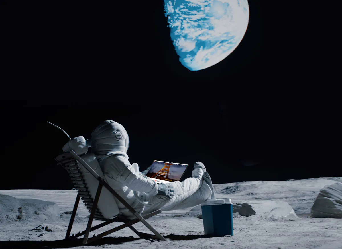 Ein Astronaut sitzt entspannt in einem Liegestuhl auf der grauen Mondoberfläche und betrachtet einen Laptop auf seinem Schoß. Auf dem Bildschirm ist eine Website mit dem Förderturm der Zeche Zollverein zu sehen. Rechts neben dem Stuhl steht eine blaue Kühlbox. Im Hintergrund leuchtet die Erde groß und hell am schwarzen Himmel. Die Szene wirkt surreal und humorvoll, als Verbindung von Weltraum, Technologie und Industriekultur.