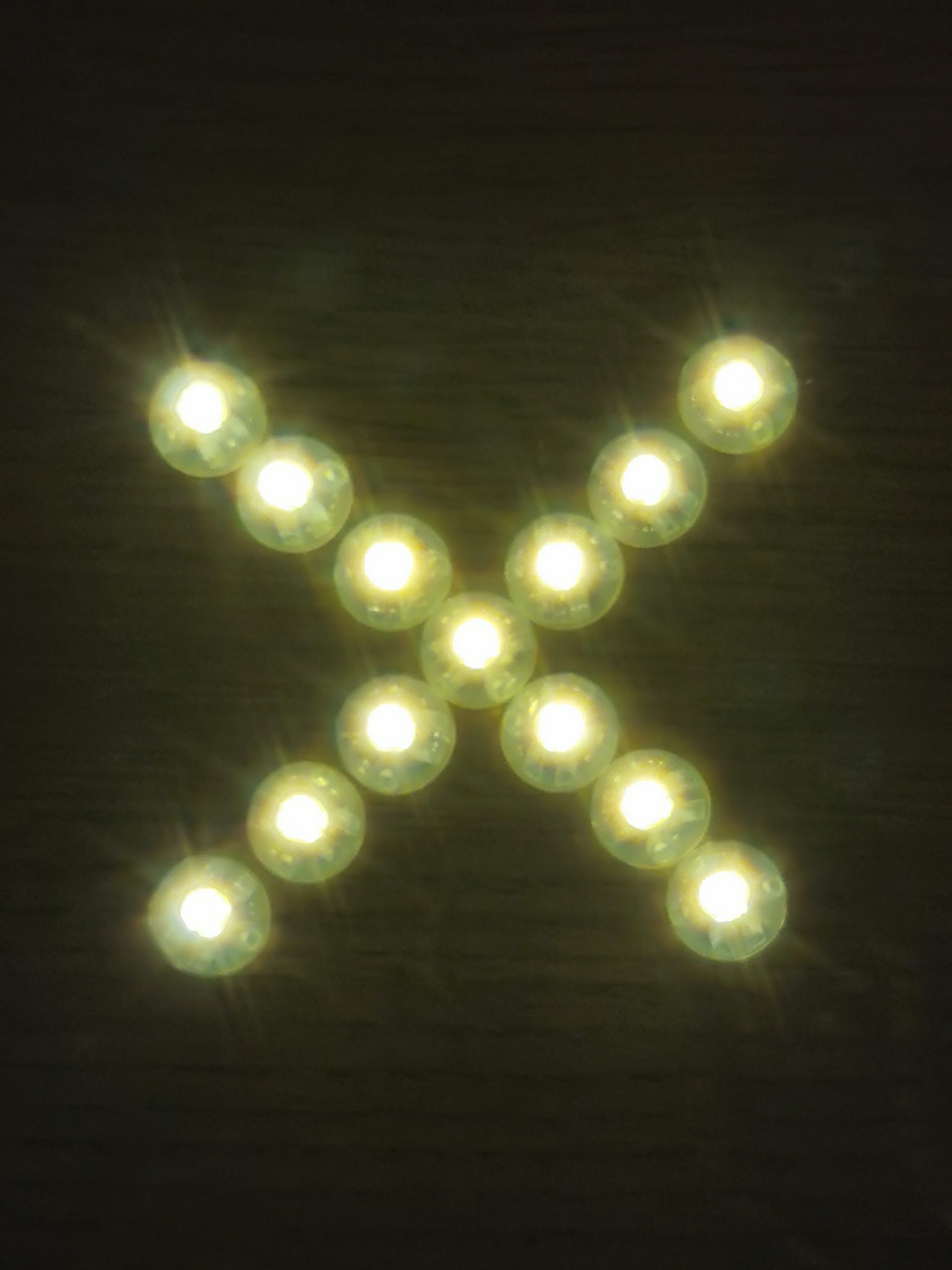 Dreizehn kleine, helle LED-Lichter sind auf einer dunklen Oberfläche in Form eines X angeordnet. Jedes Licht strahlt ein warmweißes Leuchten aus, das sternförmig in alle Richtungen abstrahlt. Der Hintergrund ist dunkelbraun und leicht strukturiert, vermutlich Holz. Die Anordnung der Lichter ergibt eine symmetrische, diagonale Kreuzform.