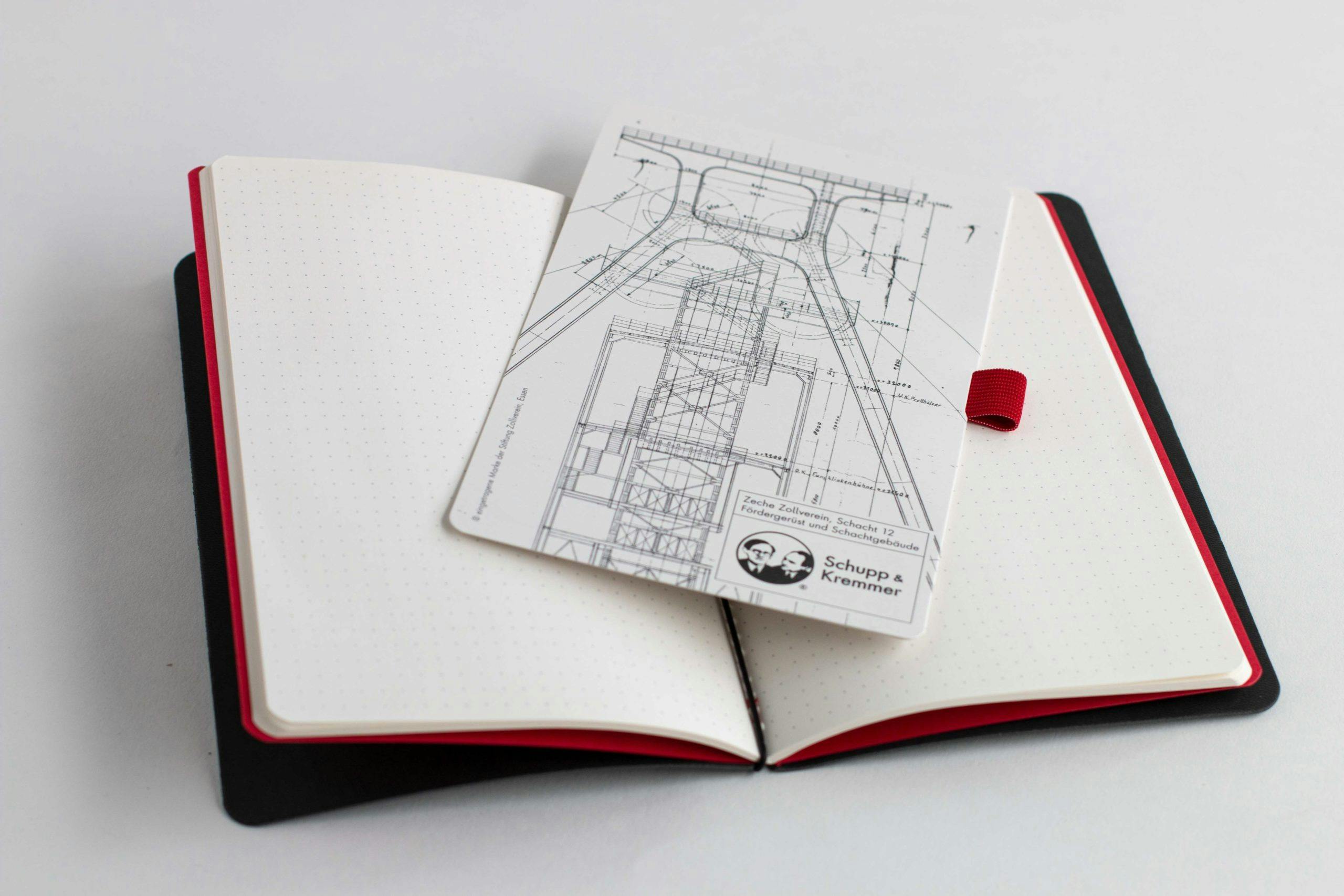 Ein geöffnetes Notizbuch mit schwarzem Einband und rotem Rand liegt auf einer weißen Fläche. Auf den punktkarierten Seiten liegt eine Postkarte oder ein Plan mit einer technischen Zeichnung des Fördergerüsts der Zeche Zollverein, Schacht 12. Unten rechts auf der Karte befindet sich ein Logo mit den Porträts der Architekten Schupp und Kremmer. Die Szene wirkt aufgeräumt und modern, mit Bezug auf Architektur und Industriekultur.