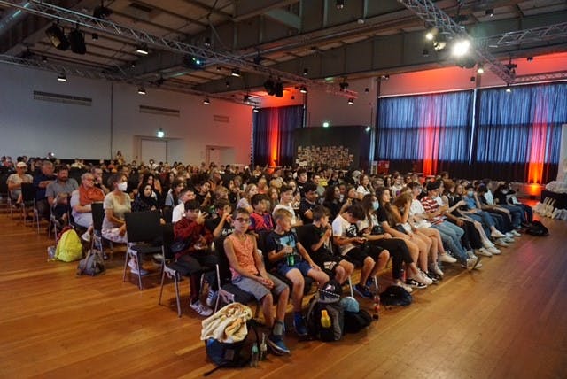 Großer Veranstaltungsraum mit hoher Decke und Beleuchtungstechnik, in dem viele Menschen, überwiegend Kinder und Jugendliche, auf Stuhlreihen sitzen und aufmerksam zur Bühne blicken. Einige klatschen, andere tragen Rucksäcke oder halten Trinkflaschen. Im Hintergrund sitzen auch Erwachsene, teils mit Gesichtsmasken. Der Saal ist stimmungsvoll mit rotem Licht an den Seitenwänden beleuchtet, die Fenster sind mit dunklen Vorhängen verdeckt. Die Atmosphäre wirkt lebendig und gespannt, als handele es sich um eine Präsentation oder Abschlussfeier.