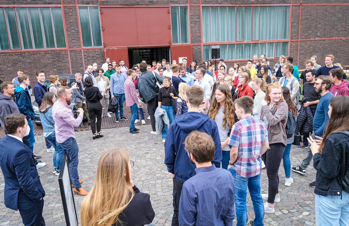 Eine große Gruppe junger Menschen steht auf einem gepflasterten Platz vor einem Backsteingebäude mit roten Türen und Fenstern. Viele unterhalten sich, hören zu oder schauen sich um. Einige tragen Rucksäcke, Jacken oder Mappen. Die Atmosphäre wirkt lebendig und interessiert, als handele es sich um eine Einführungsveranstaltung, eine Führung oder ein Gruppenfoto im Rahmen eines Workshops oder Bildungsevents. Im Hintergrund ist eine geöffnete Tür zu sehen, aus der weitere Personen kommen. Die Szene vermittelt Gemeinschaft und Aufbruchsstimmung.