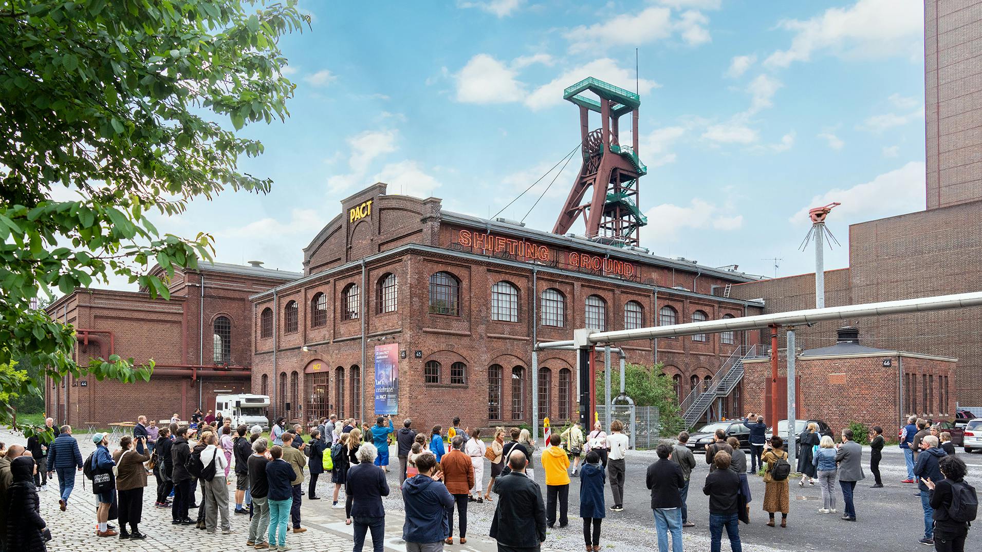 Vor dem historischen Backsteingebäude des PACT Zollverein auf dem Gelände der Zeche Zollverein versammelt sich eine größere Gruppe von Menschen. Das Gebäude trägt in großen leuchtenden Buchstaben den Schriftzug Shifting Ground. Im Hintergrund ragt ein Fördergerüst mit seiner roten Stahlkonstruktion in den Himmel. Die Szenerie spielt bei Tageslicht unter einem leicht bewölkten Himmel. Einige Personen fotografieren oder beobachten aufmerksam das Geschehen, während andere in kleinen Gruppen stehen und sich unterhalten.