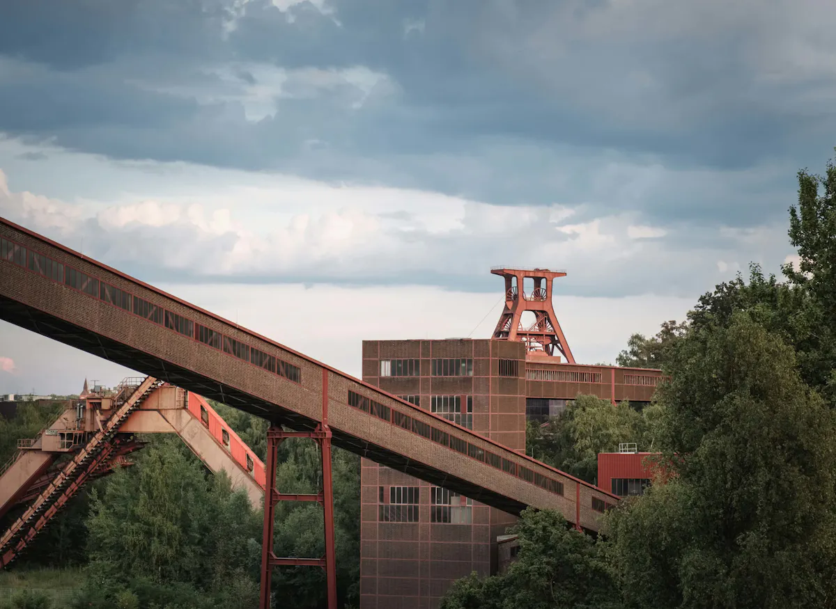 Blick auf das Welterbe Zollverein in Essen mit dem markanten Doppelbock-Fördergerüst von Schacht XII im Hintergrund. Im Vordergrund verläuft eine rote Förderbrücke schräg durch das Bild, umgeben von Bäumen und Industriearchitektur. Der Himmel ist wolkig mit blau-grauen und weißen Wolken, was der Szene eine leicht dramatische Stimmung verleiht.