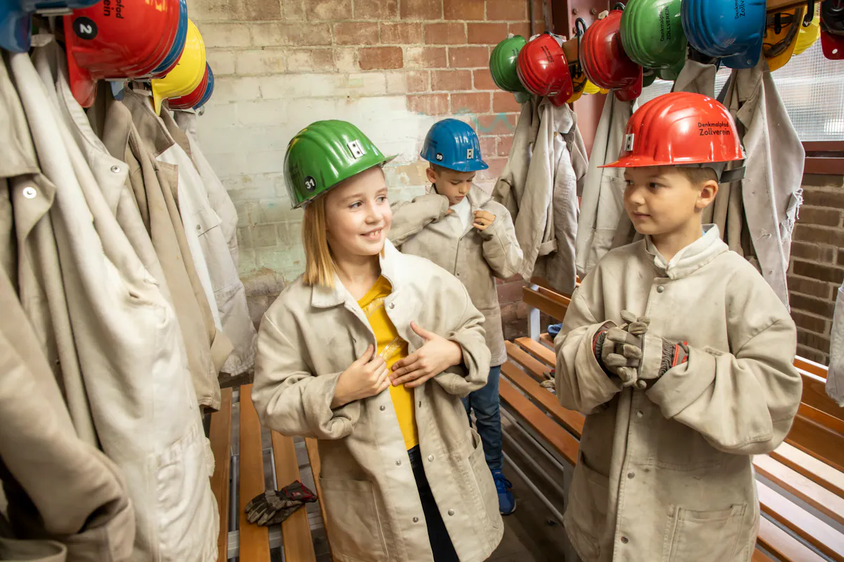 Drei Kinder stehen in einem Umkleideraum mit Backsteinwand und Holzbänken. Sie tragen helle Schutzjacken und bunte Schutzhelme in Rot, Grün und Blau. Zwei Kinder unterhalten sich lächelnd, während das dritte im Hintergrund seine Jacke schließt. An der Wand hängen weitere Helme und Jacken ordentlich an Haken. Die Szene vermittelt den Eindruck einer Vorbereitung auf eine kindgerechte Führung oder Aktivität in einem industriekulturellen Lernort wie der Zeche Zollverein.