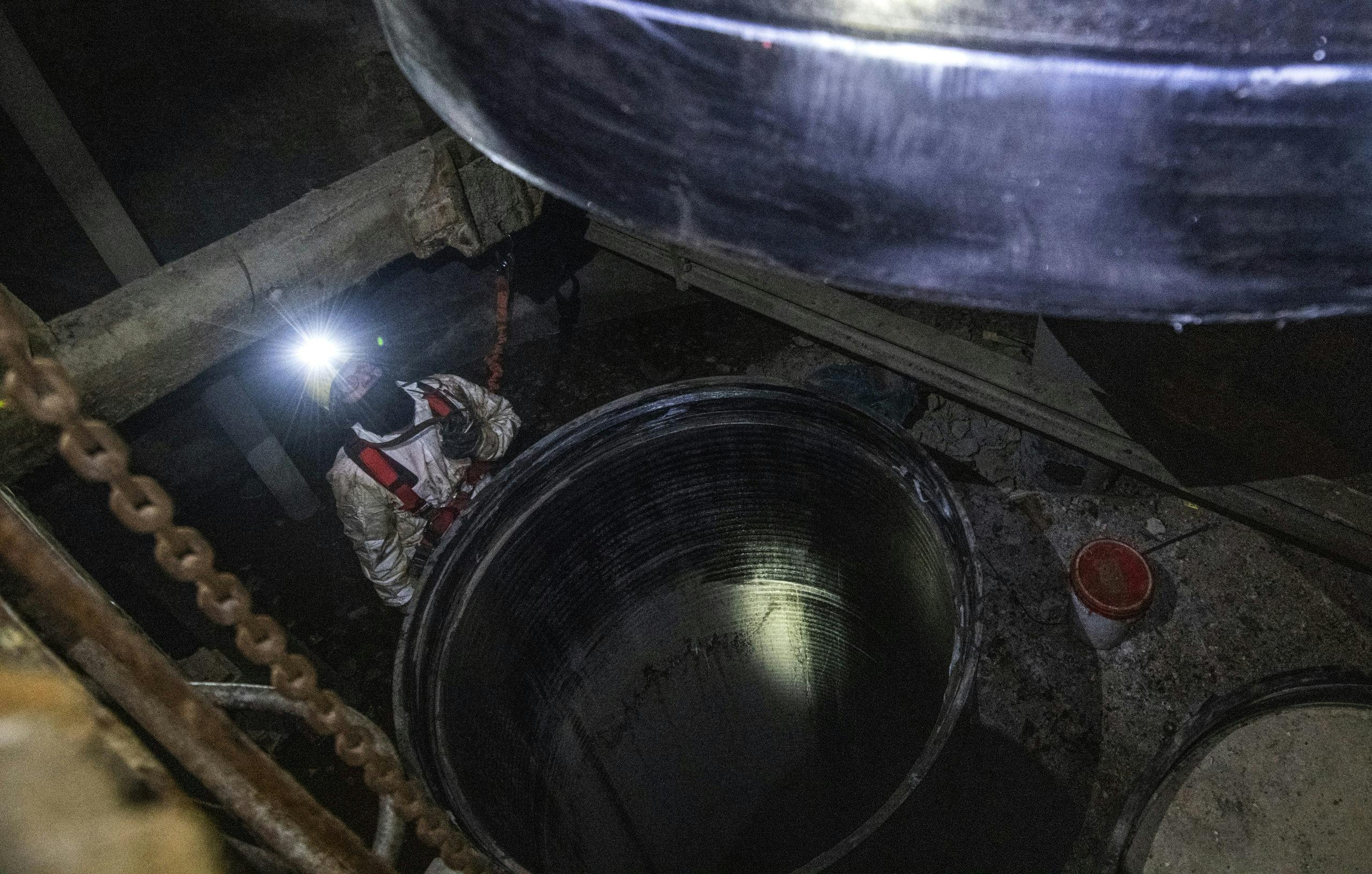 Eine Person in Schutzkleidung mit Helm und Stirnlampe steht in einem dunklen, industriellen Raum vor einer großen, offenen Röhre oder einem Schacht. Der Lichtkegel der Stirnlampe leuchtet auf die Innenseite der Röhre. Um die Person herum liegen Rohre, Ketten und Werkzeuge auf dem staubigen Boden. Die Szene wirkt wie ein technischer Einsatz in einem stillgelegten Industriegebäude oder einer Anlage zur Wartung oder Inspektion.