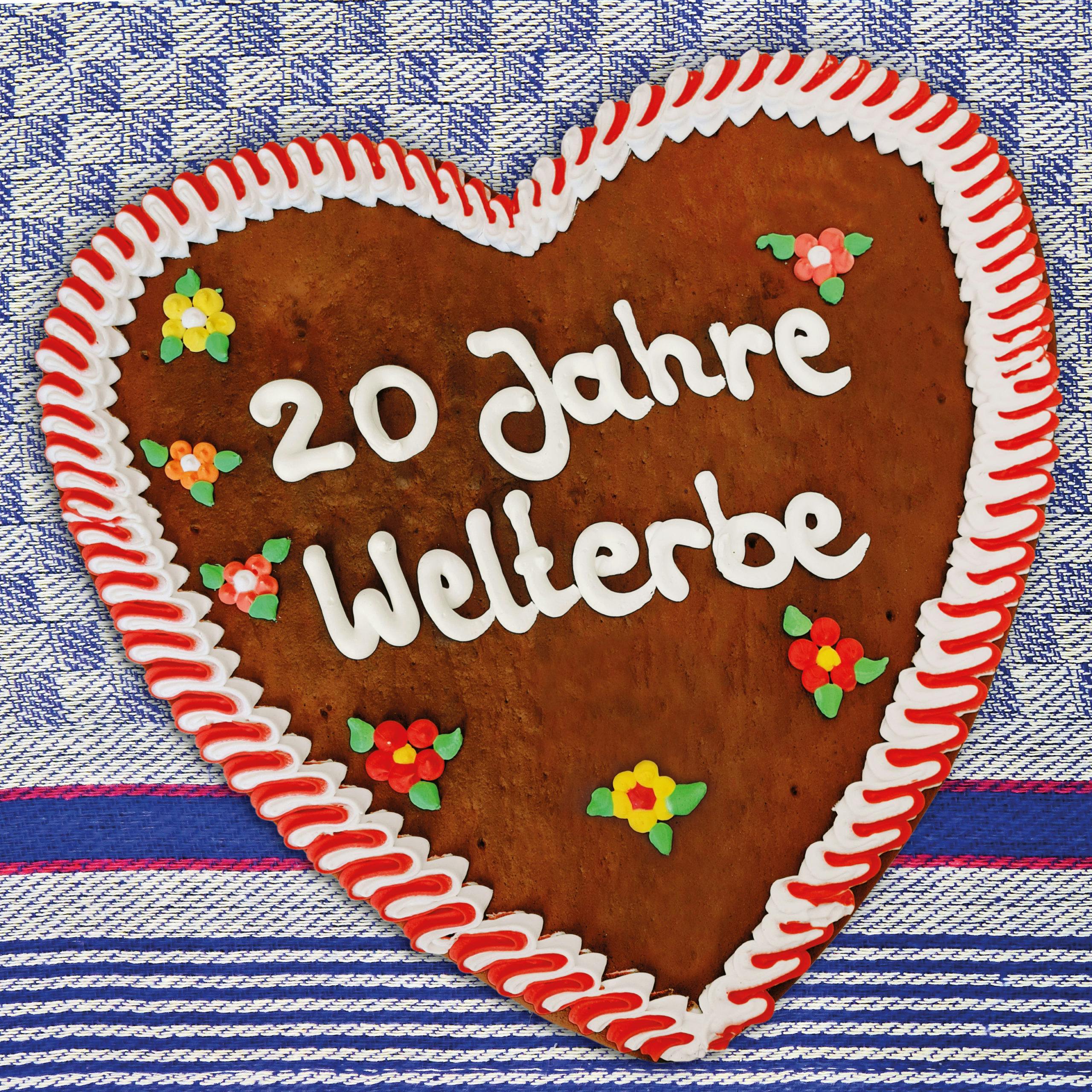 Ein herzförmiges Lebkuchenherz liegt auf einer blau-weiß karierten Stoffunterlage. Es ist mit weißem und rotem Zuckerguss am Rand verziert und trägt in weißer Zuckerschrift die Aufschrift 20 Jahre Welterbe. Um den Schriftzug herum sind kleine bunte Zuckerdekoblumen in Rot, Gelb und Orange verteilt.